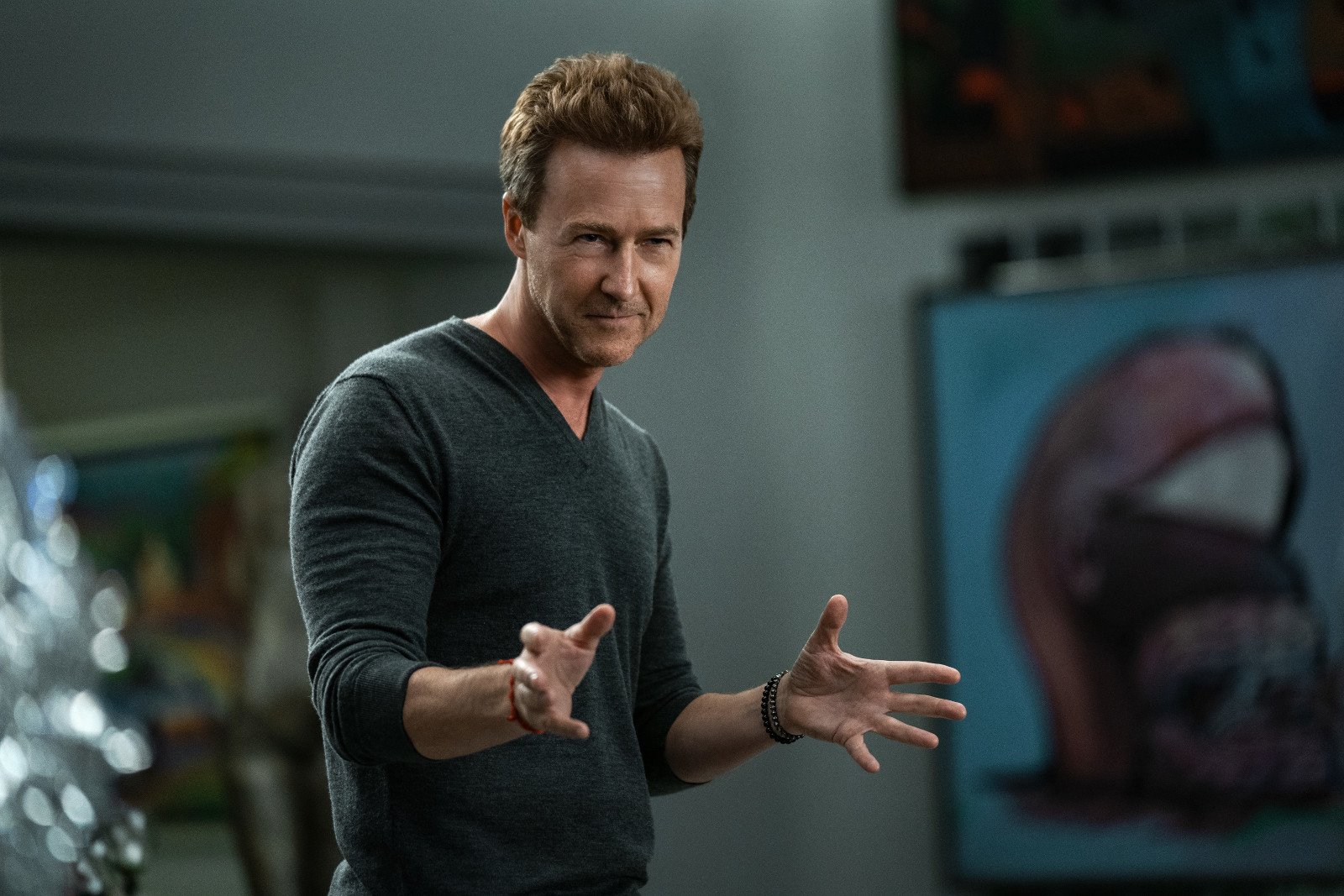 Edward Norton as Miles Bron in 'Glass Onion' for our article about its ending. He's wearing a grey, long-sleeved shirt and holding his hands out.