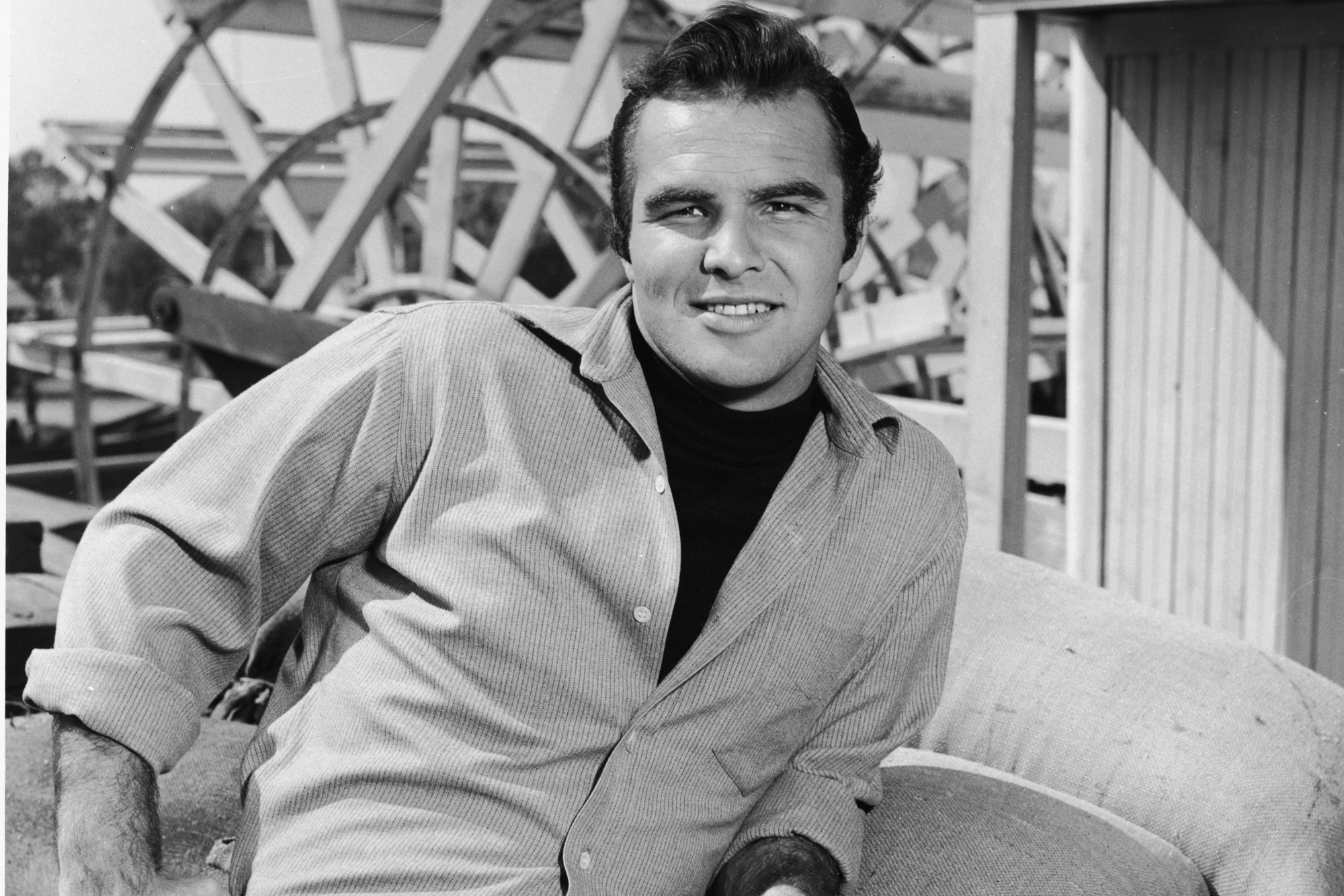 'Gunsmoke' actor Burt Reynolds on 'Riverboat' set smiling in a button-up shirt and leaning on sandbags