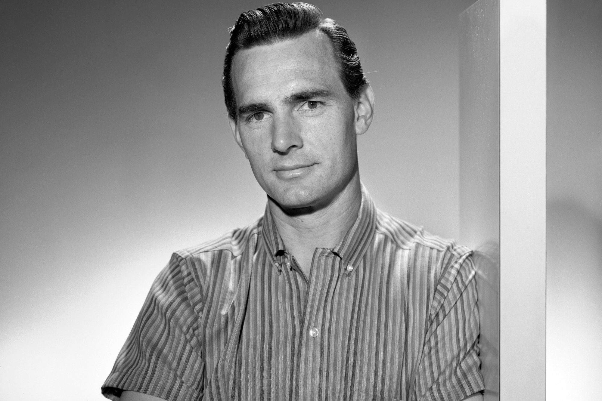 'Gunsmoke' actor Dennis Weaver in a black-and-white picture with his arms crossed wearing a striped collared-shirt