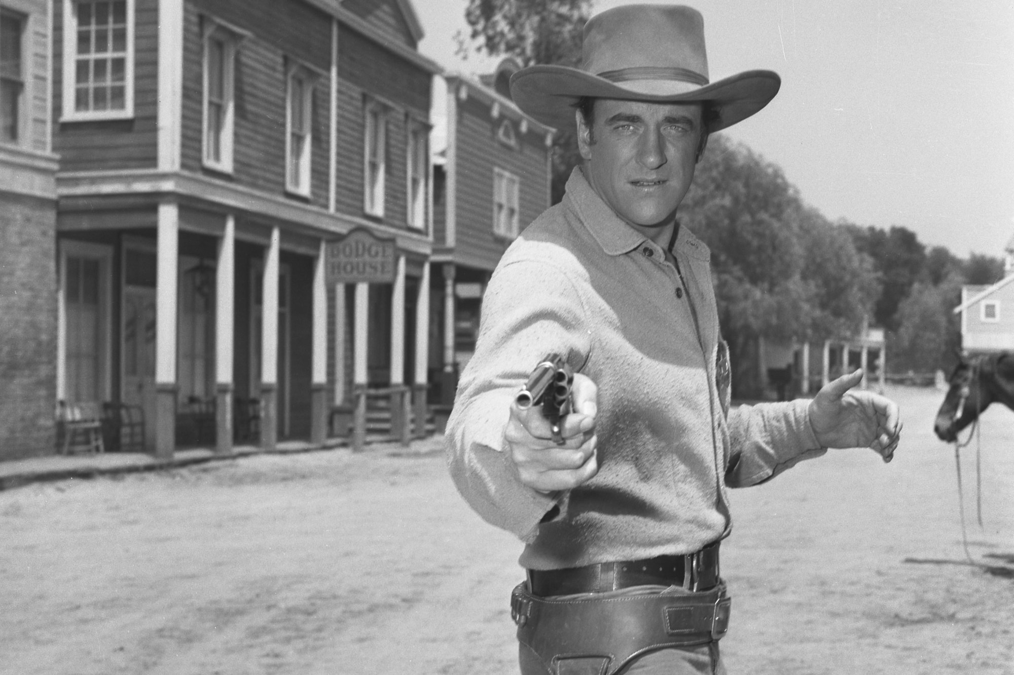 'Gunsmoke' actor James Arness as Matt Dillon in a black-and-white picture on a dirt road, wearing Western clothes, and holding a gun out at the camera