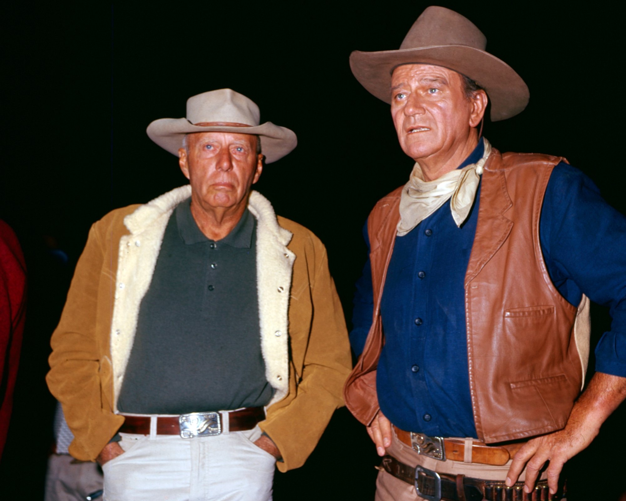 Howard Hawks and John Wayne wearing Western outfits, looking surprised in front of a dark background