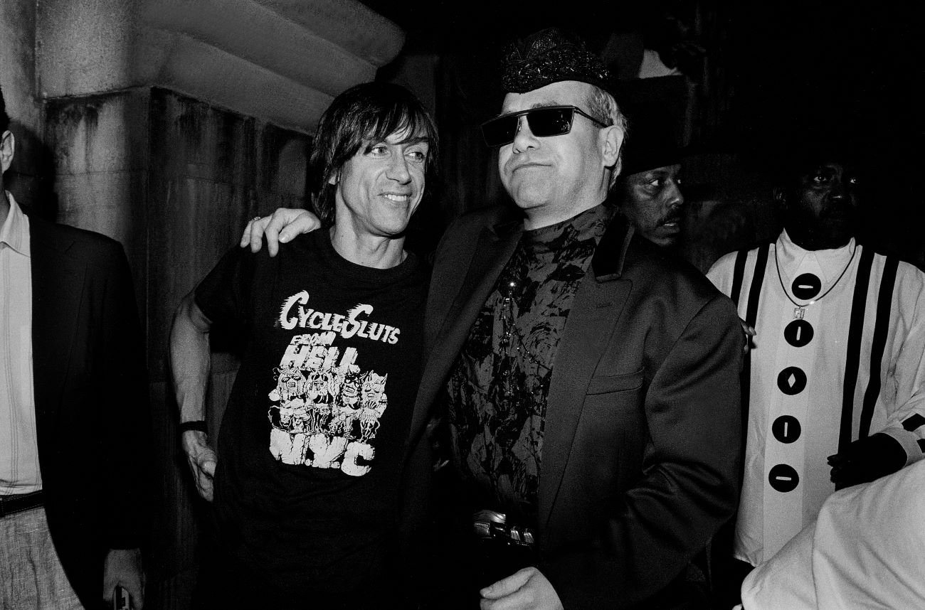 A black and white photo of Elton John with his arm around Iggy Pop's shoulders.