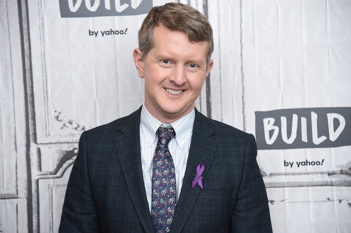 'Jeopardy!' host Ken Jennings wearing a grey suit and purple tie; smiles for a photo.