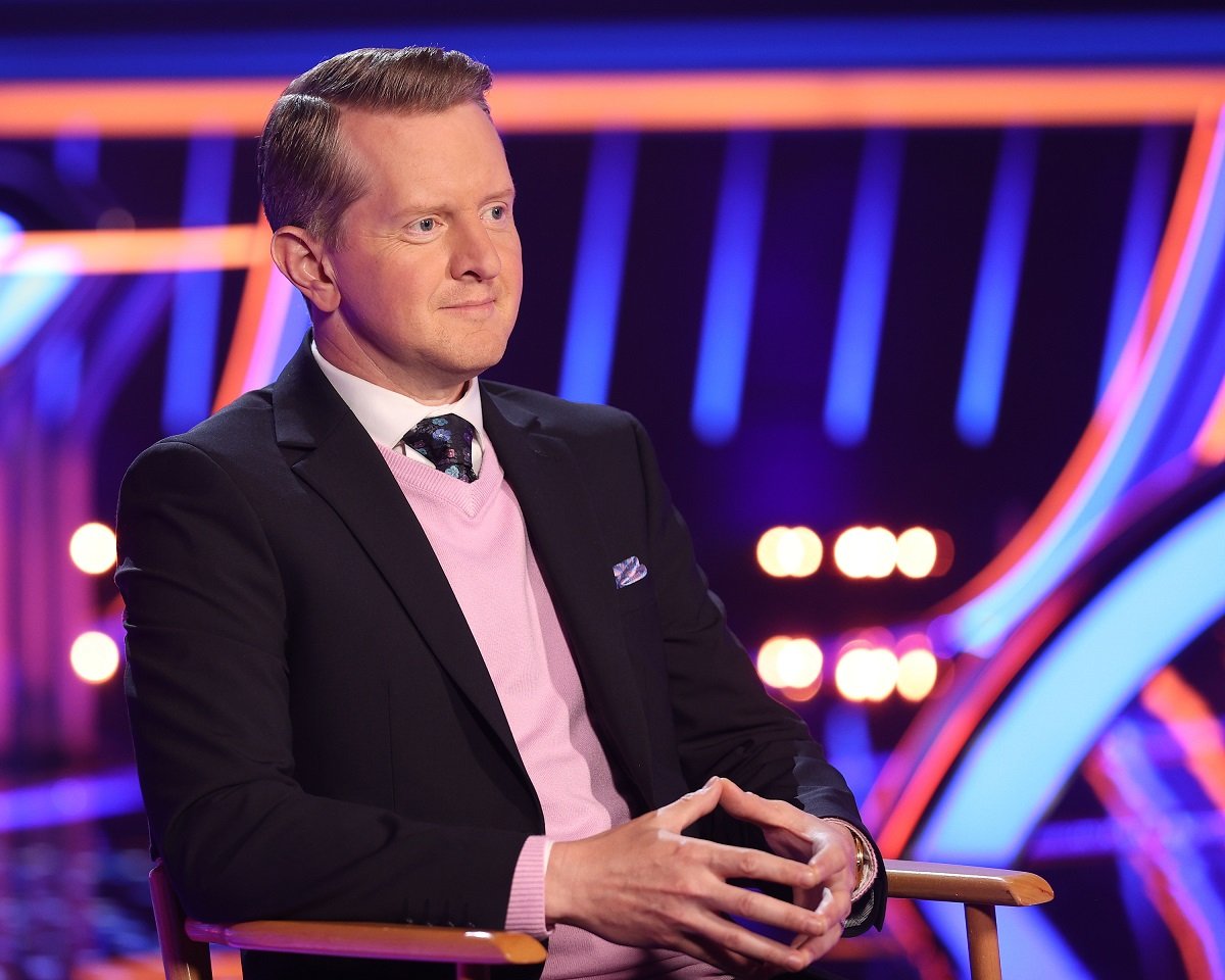 'Jeopardy' host Ken Jennings wearing a black suit and pink vest; sits in a director's chair.
