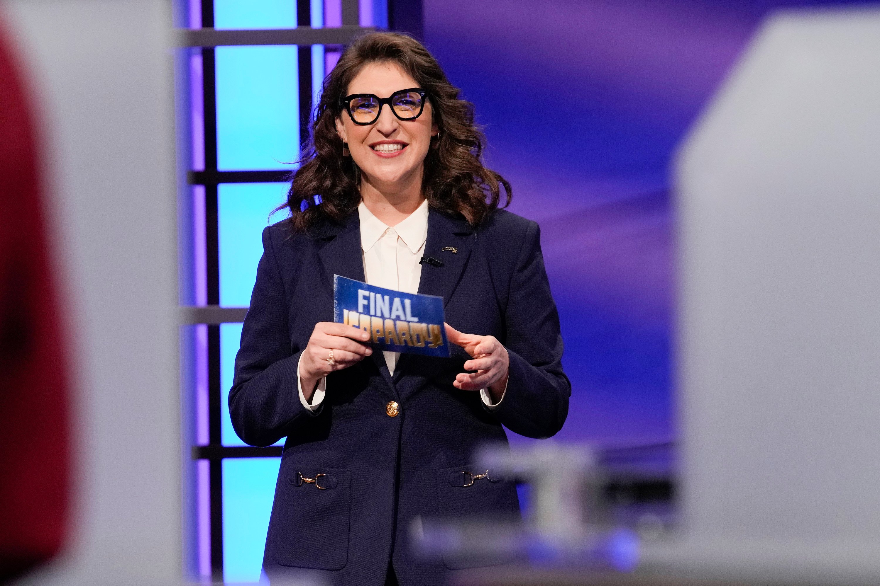 'Jeopardy!' host Mayim Bialik wearing glasses and a navy blue suit while filming an episode of the game show.