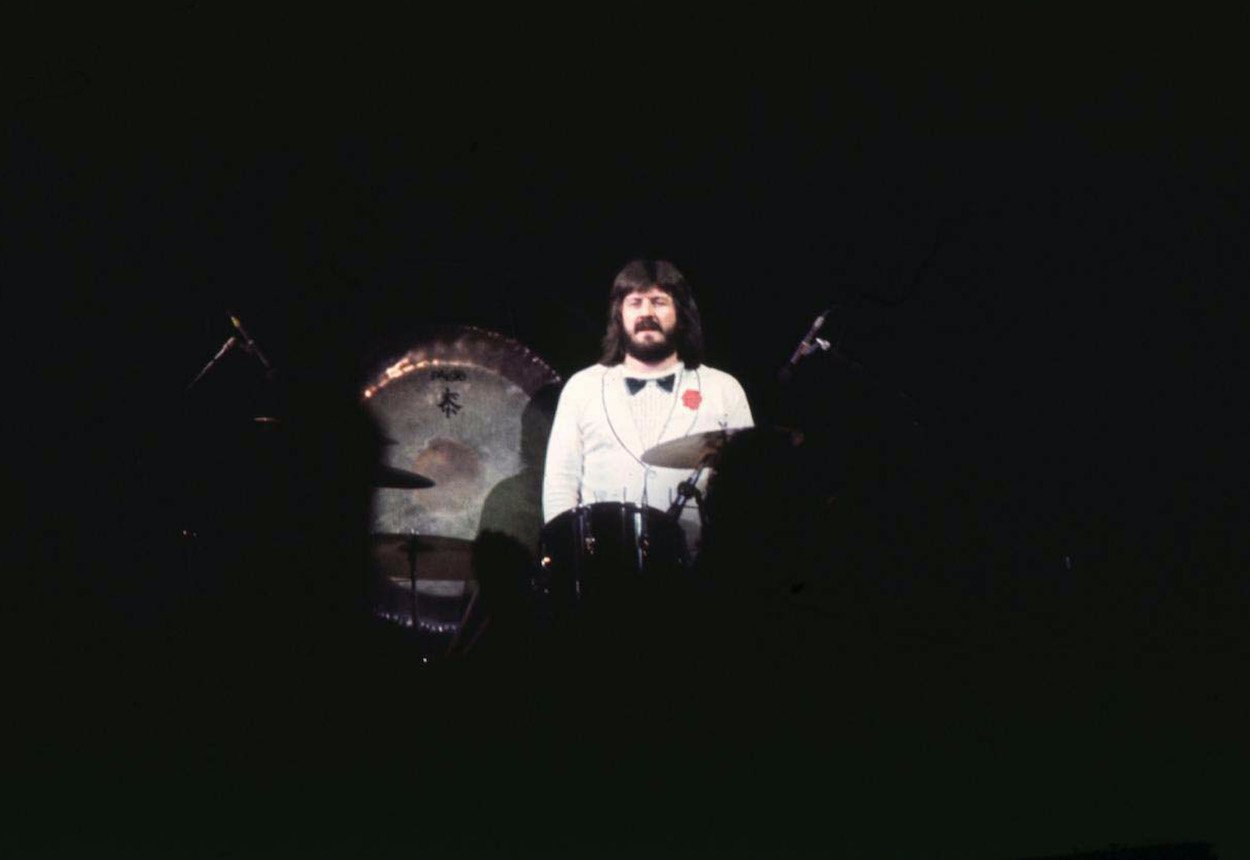 John Bonham stands behind his drum kit wearing a faux white tuxedo during a 1977 Led Zeppelin concert.