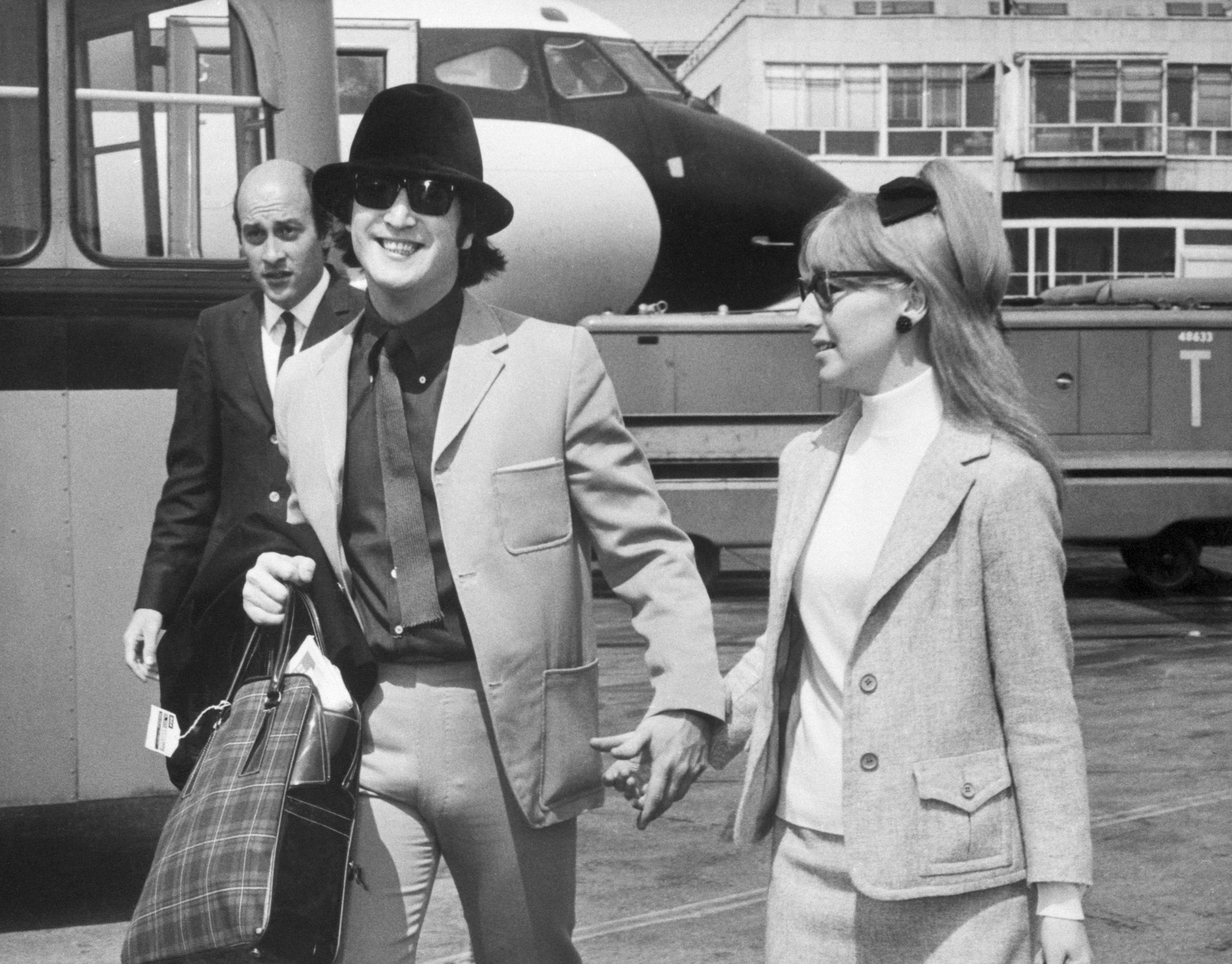 A black and white picture of John Lennon and Cynthia Lennon holding hands at the airport.