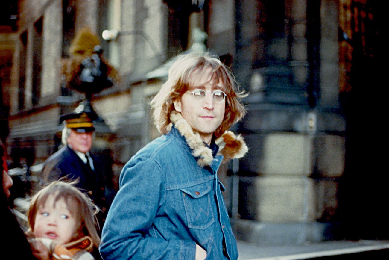 John Lennon wears a denim vest with a fur collar and walks outside.