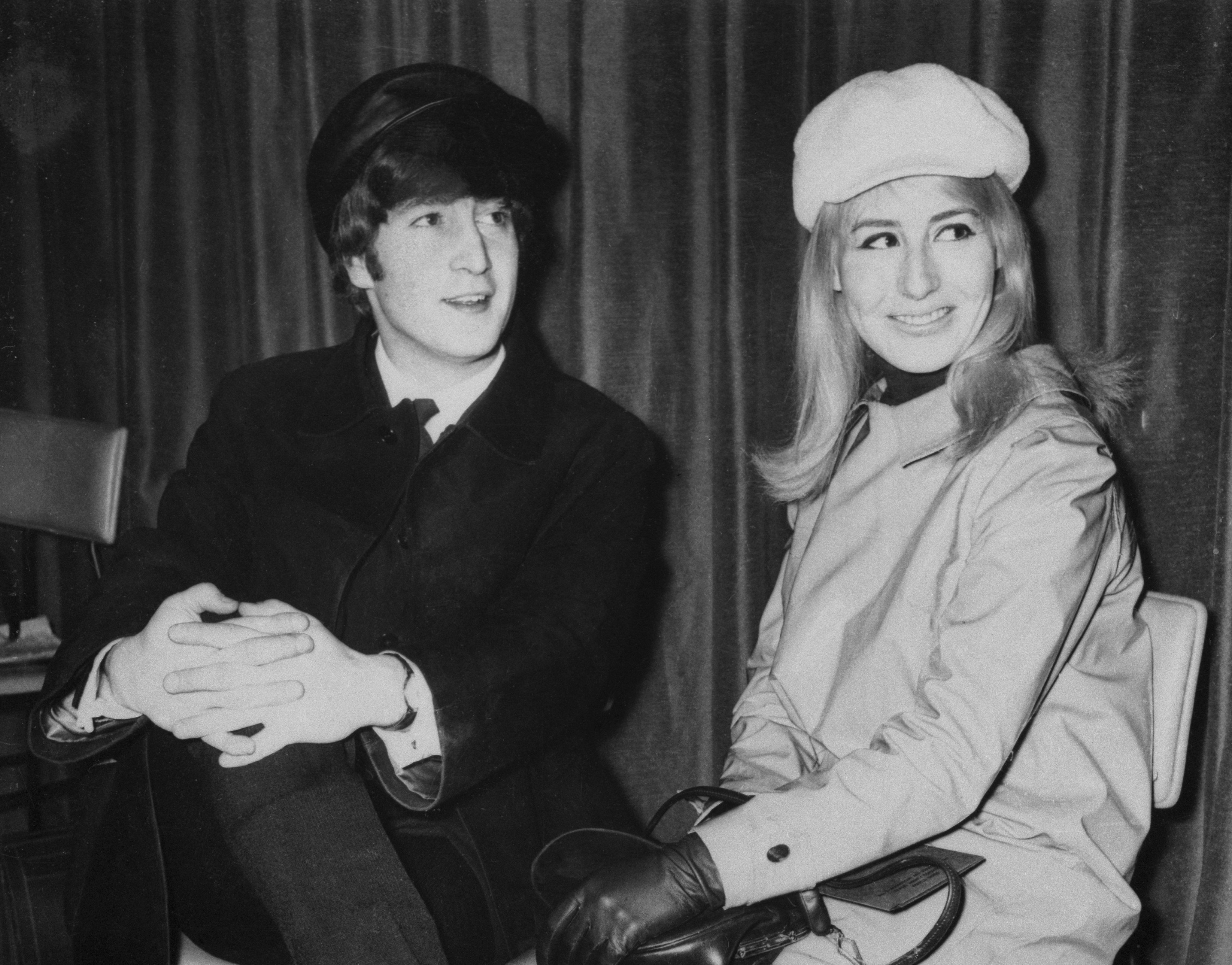 A black and white picture of John Lennon and his wife Cynthia Lennon wearing coats and hats and sitting in chairs.