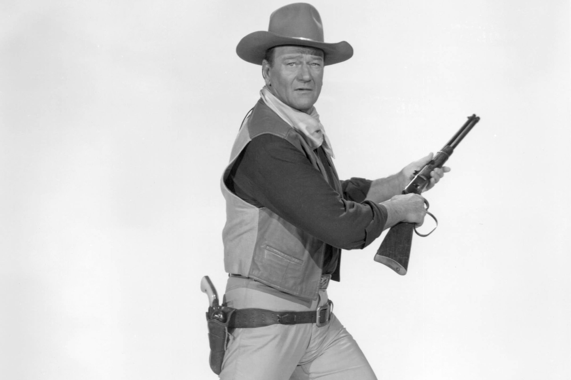 John Wayne, a movie star who made Westerns. A black-and-white picture with him wearing a cowboy costume and holding a rifle in front of a white background.
