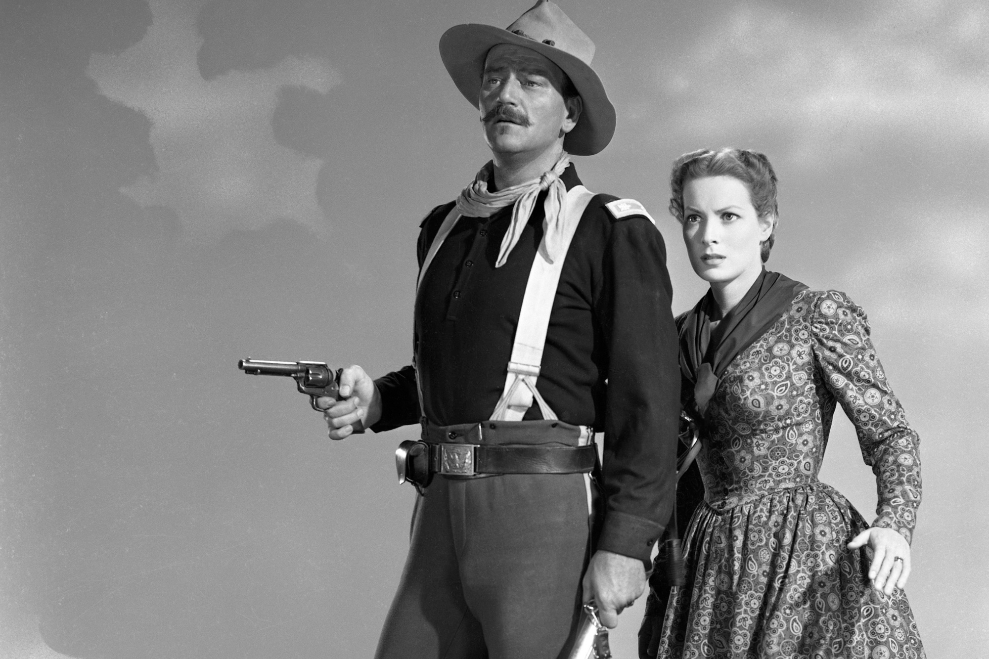 John Wayne as Lieutenant Colonel Kirby Yorke and Maureen O'Hara as Mrs Kathleen Yorke in a black-and-white picture. Wayne is holding his pistol out and O'Hara is holding onto his shoulder.