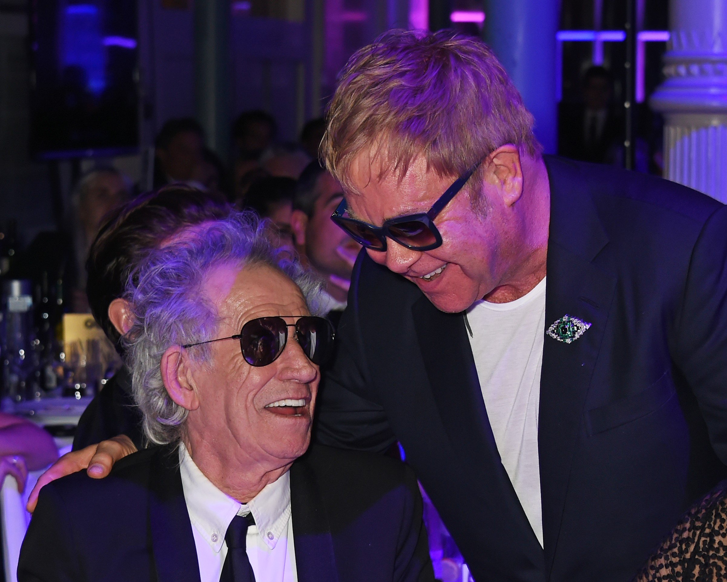 Elton John stands with his hand on Keith Richards' shoulder. They both wear sunglasses.