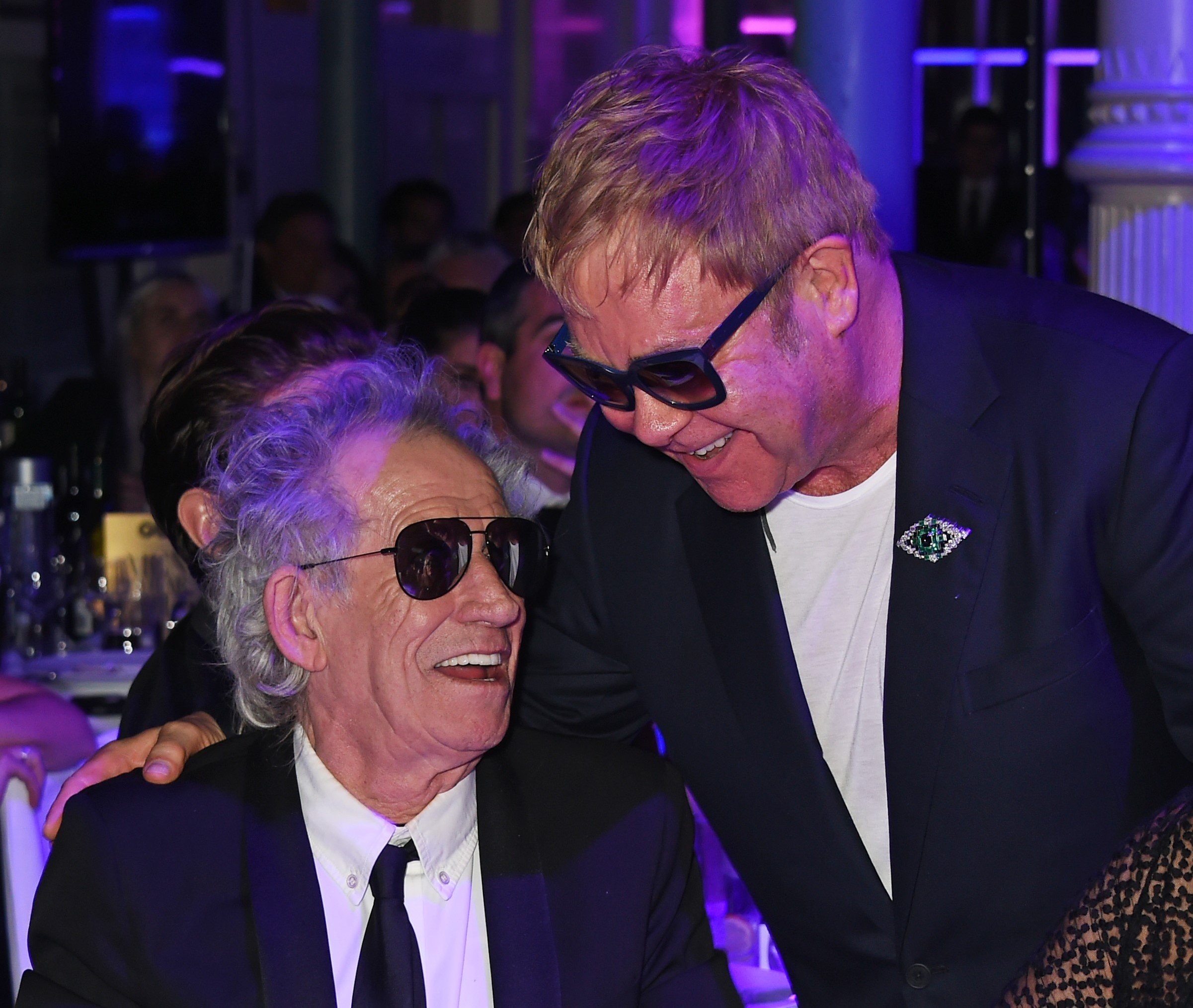 Keith Richards sits and Elton John stands with his hand on his shoulder.