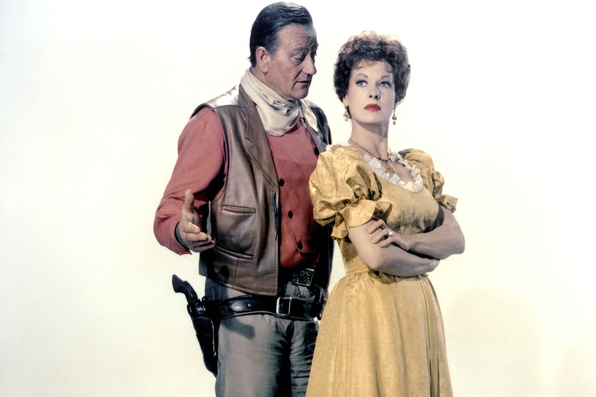 'McLintock!' actors John Wayne and Maureen O'Hara in Western costumes for a promo shoot. He's standing next to her talking, while she has her arms crossed looking away from him.