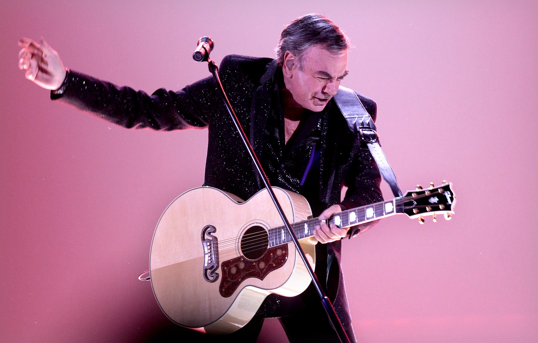 Neil Diamond playing his guitar against a pink background