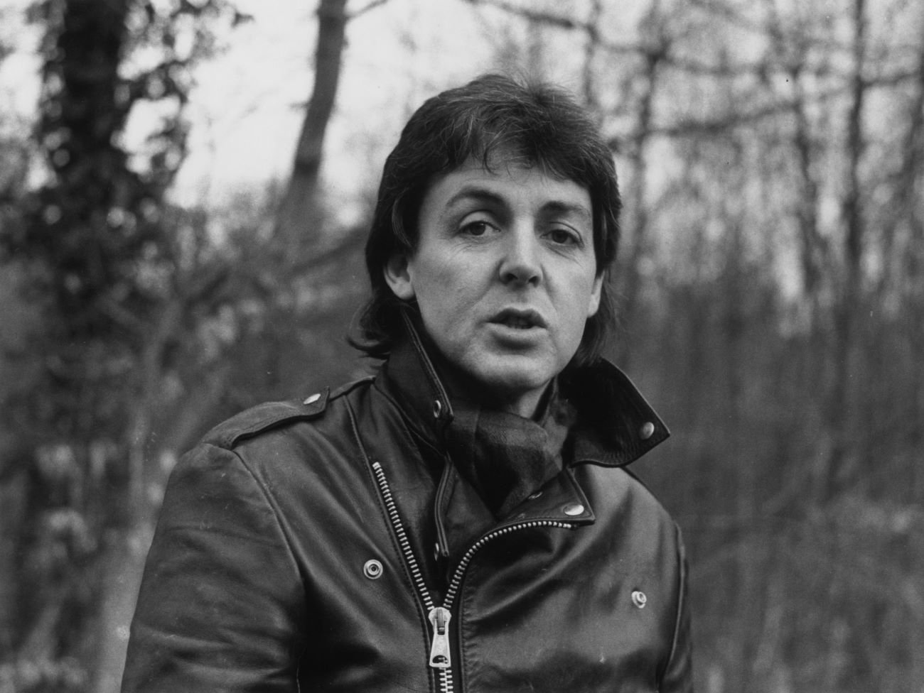 A black and white picture of Paul McCartney of The Beatles wearing a leather jacket and sitting outside. 