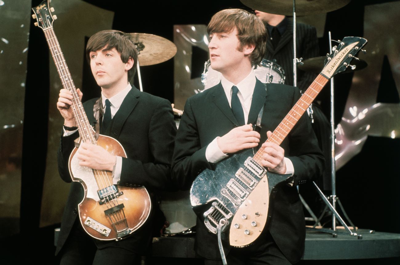 Paul McCartney and John Lennon stand onstage and hold guitars.