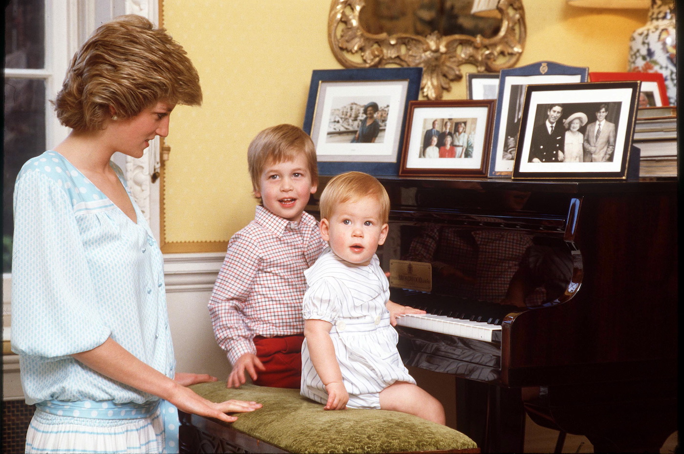 Princess Diana is nearby while Prince William and Prince Harry sit on a piano stool