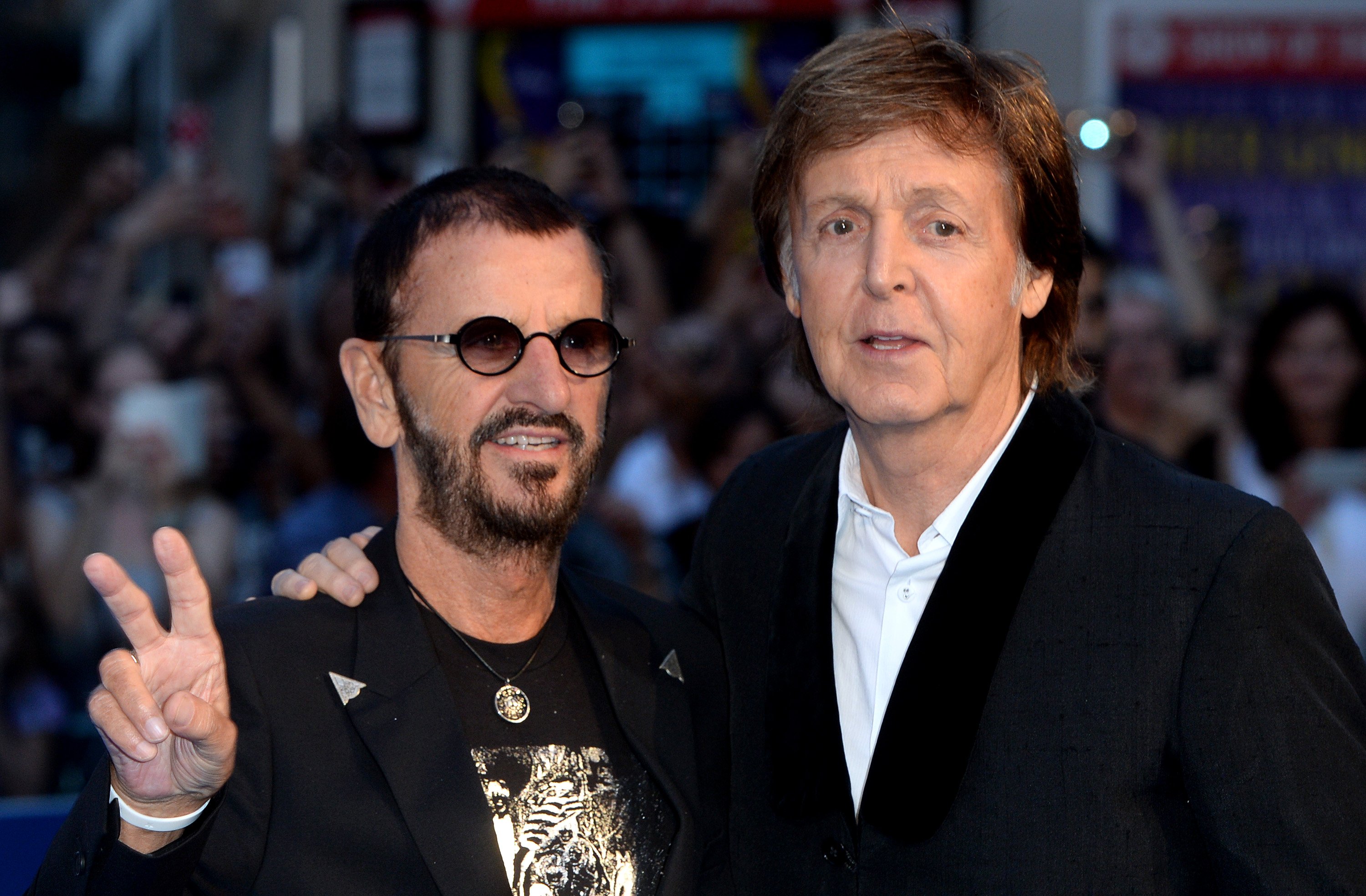Ringo Starr holds up a peace sign and Paul McCartney holds his hand around his shoulder.