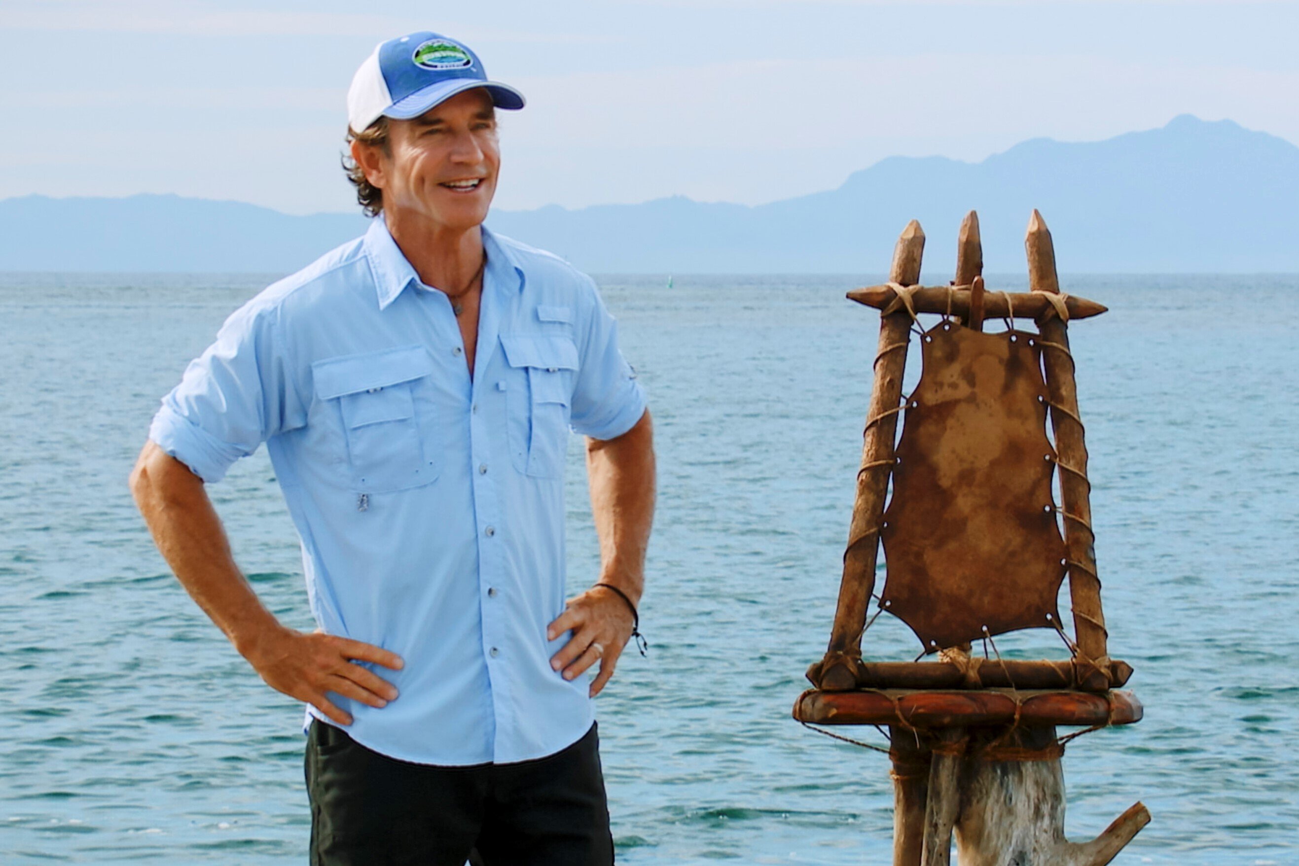 Jeff Probst, who interacts with castaway in 'Survivor' on CBS, wears a light blue button-up shirt with rolled-up sleeves, black pants, and a blue and white 'Survivor' baseball cap.