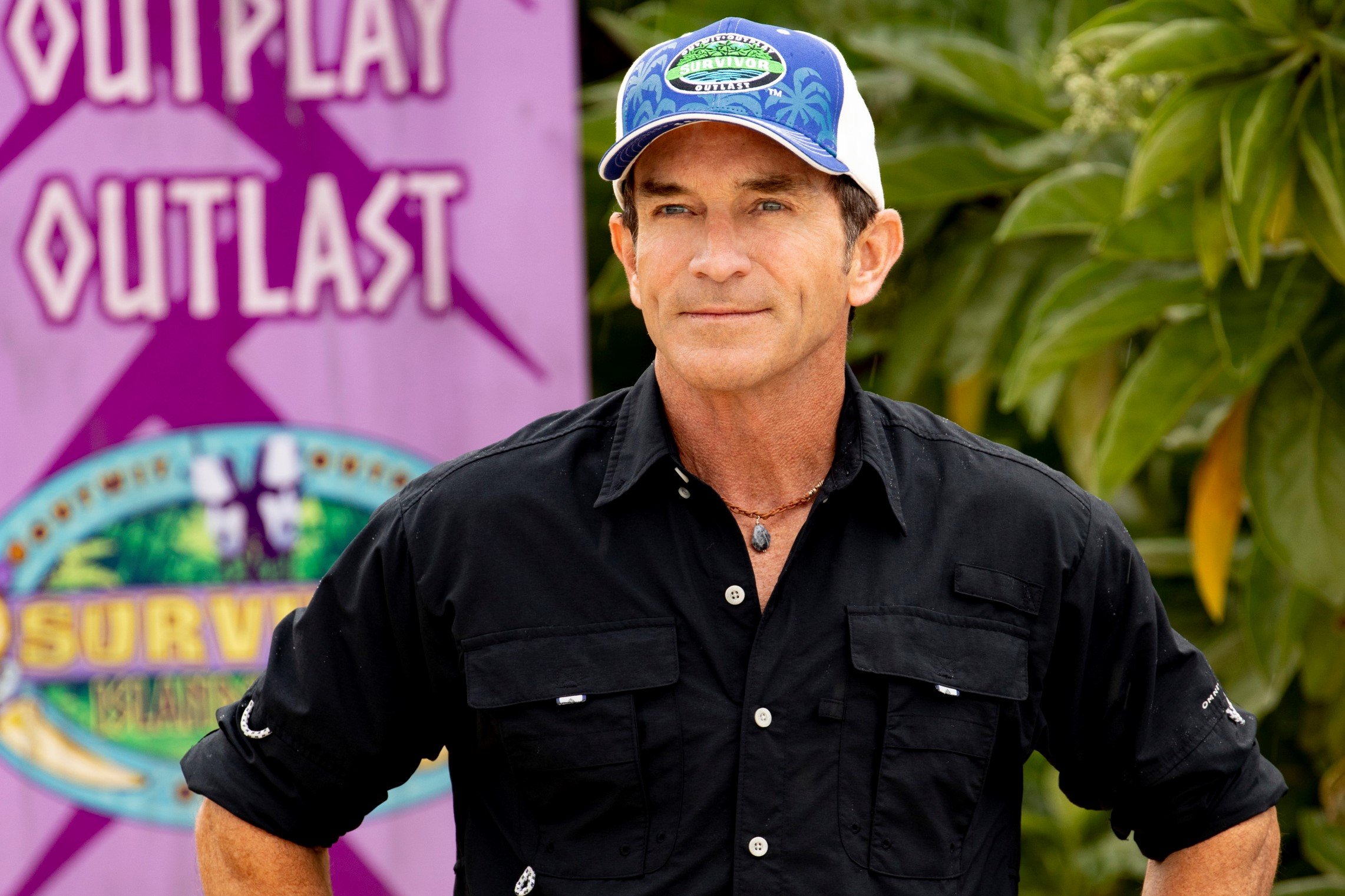 Jeff Probst, seen hosting long-running reality TV show 'Survivor' Season 39, wears a black button-up shirt rolled up at the sleeves and a blue, green, and white 'Survivor' baseball cap.