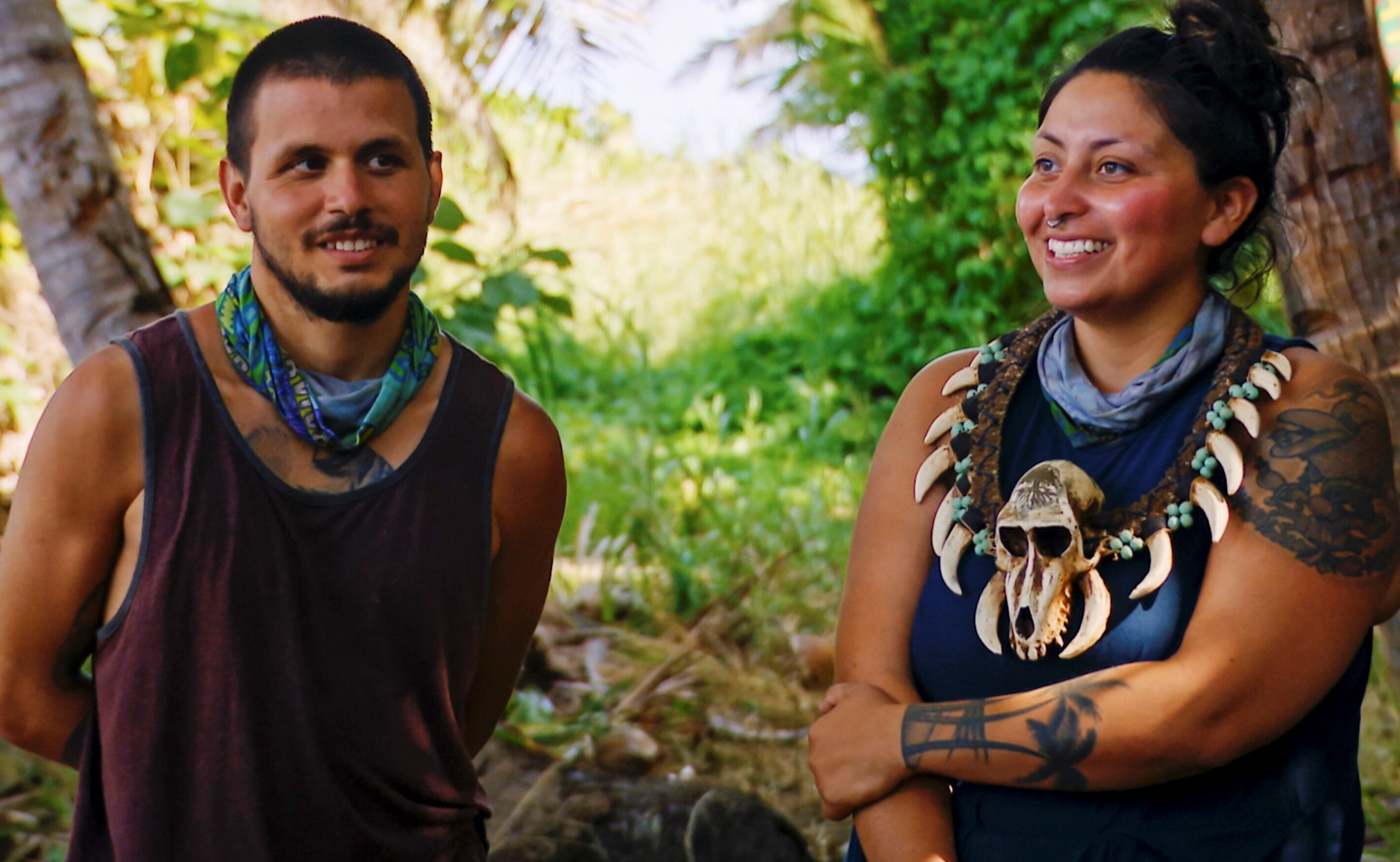 Jesse Lopez and Karla Cruz Godoy, who, according to 'Survivor' spoilers, may be headed home in the next season 43 episode, converse at camp. Jesse wears a dark red tank top and his light blue 'Survivor' buff around his neck. Karla wears a dark blue tank top, her light blue 'Survivor' buff around her neck, and the immunity necklace.