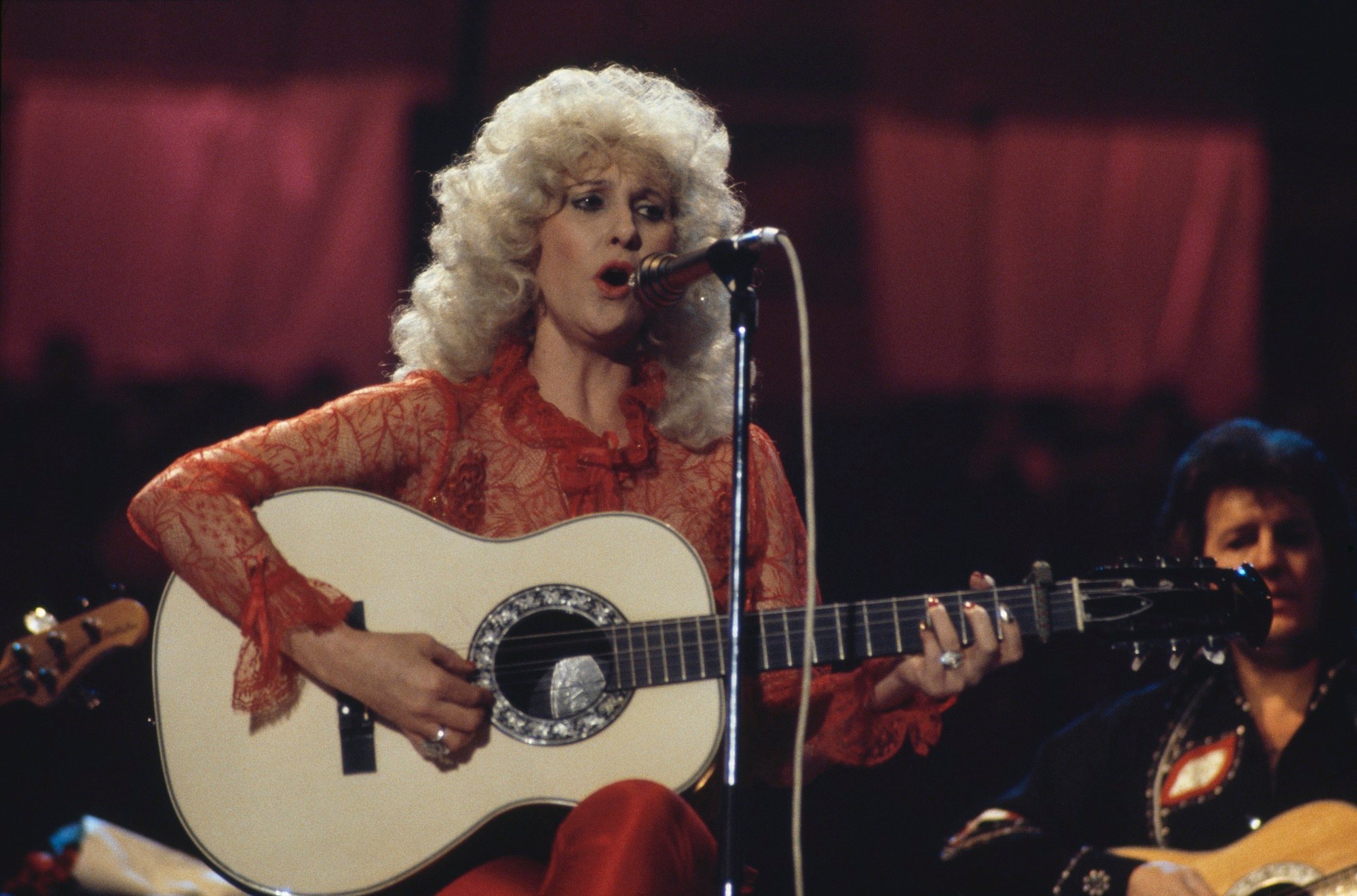 Tammy Wynette sings into a microphone while playing guitar onstage