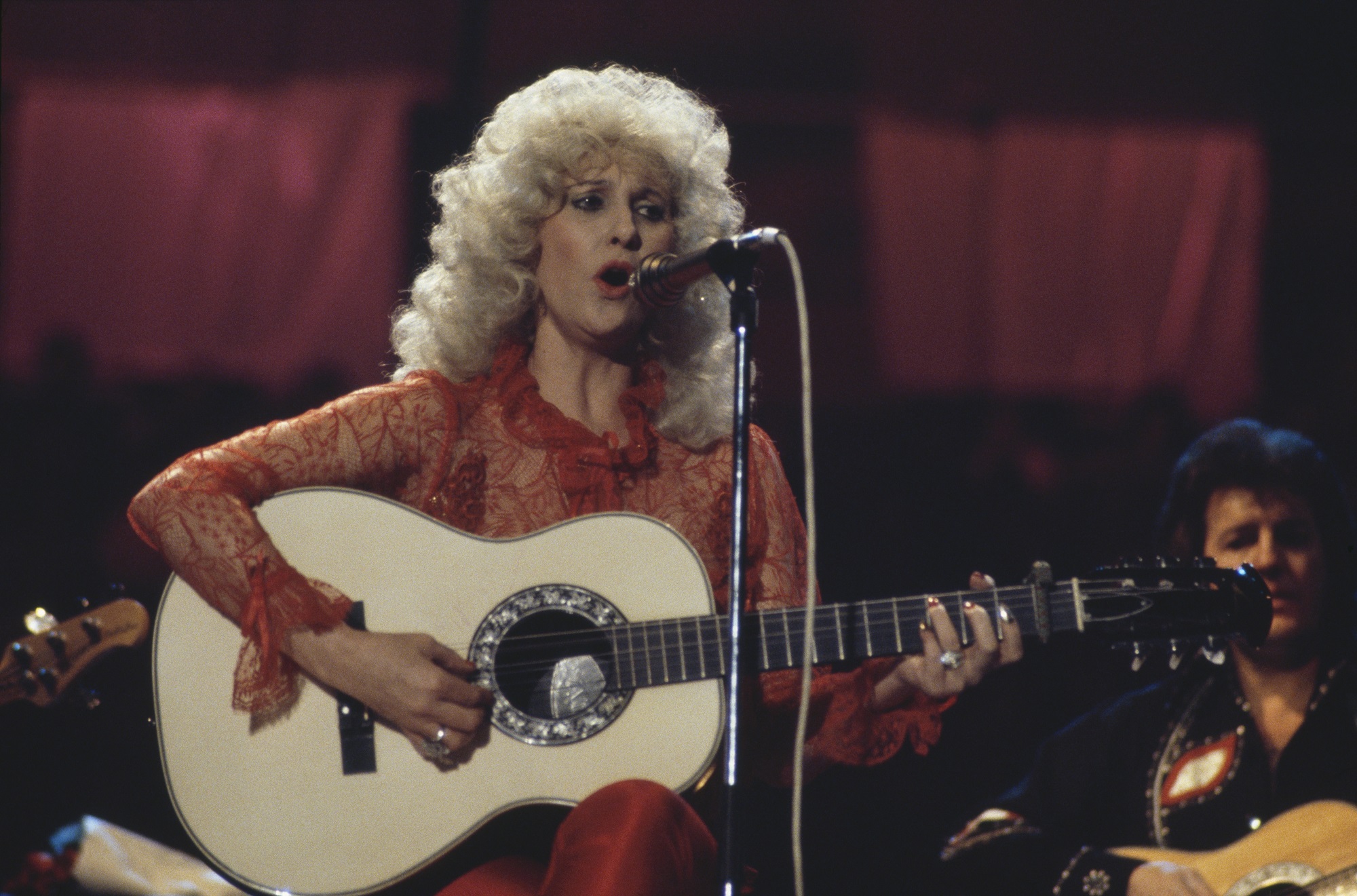 Tammy Wynette sings into a microphone while playing guitar