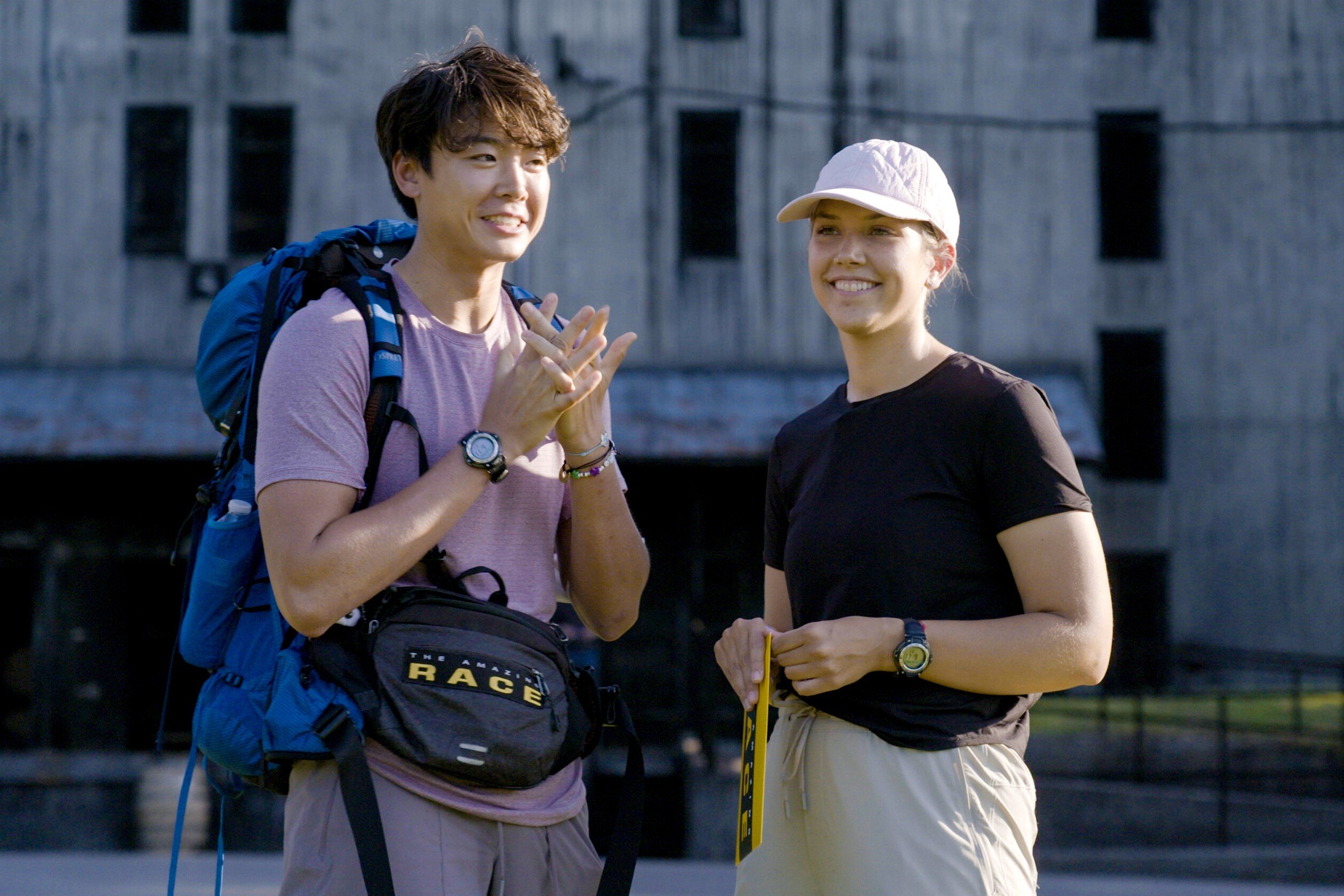 Derek Xiao and Claire Rehfuss, who, according to spoilers, made it to the finale of 'The Amazing Race 34' on CBS, read their first clue in the last leg. Derek wears a light purple shirt, light gray pants, a blue backpack, and a black 'The Amazing Race' fanny pack. Claire wears a black shirt, light gray pants, and a light pink baseball cap.