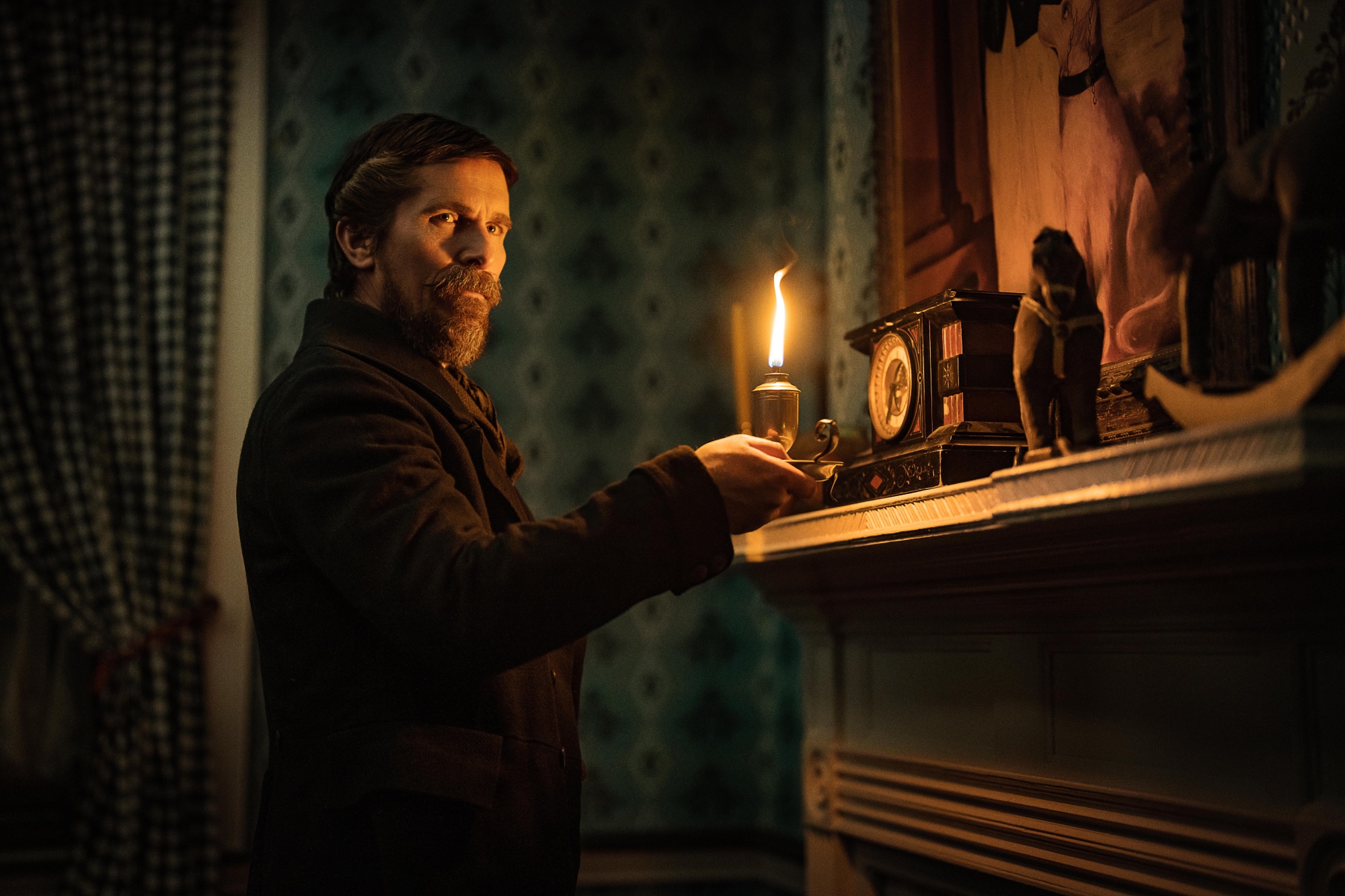 'The Pale Blue Eye' Christian Bale as Augustus Landor looking to the side, holding a candle inside of a dark room at the mantle