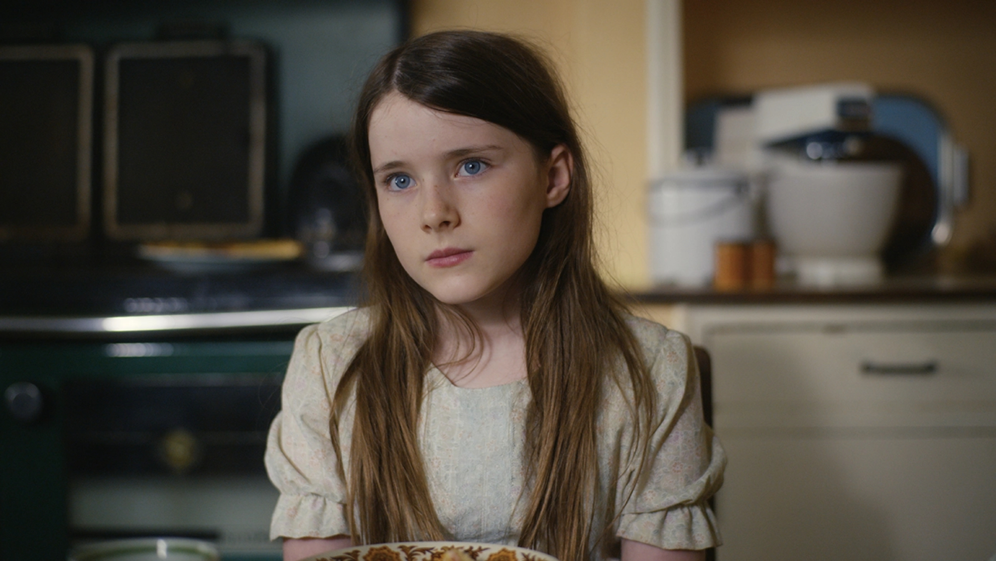 'The Quiet Girl' Catherine Clinch as Cáit sitting in a chair at the kitchen table looking unsure