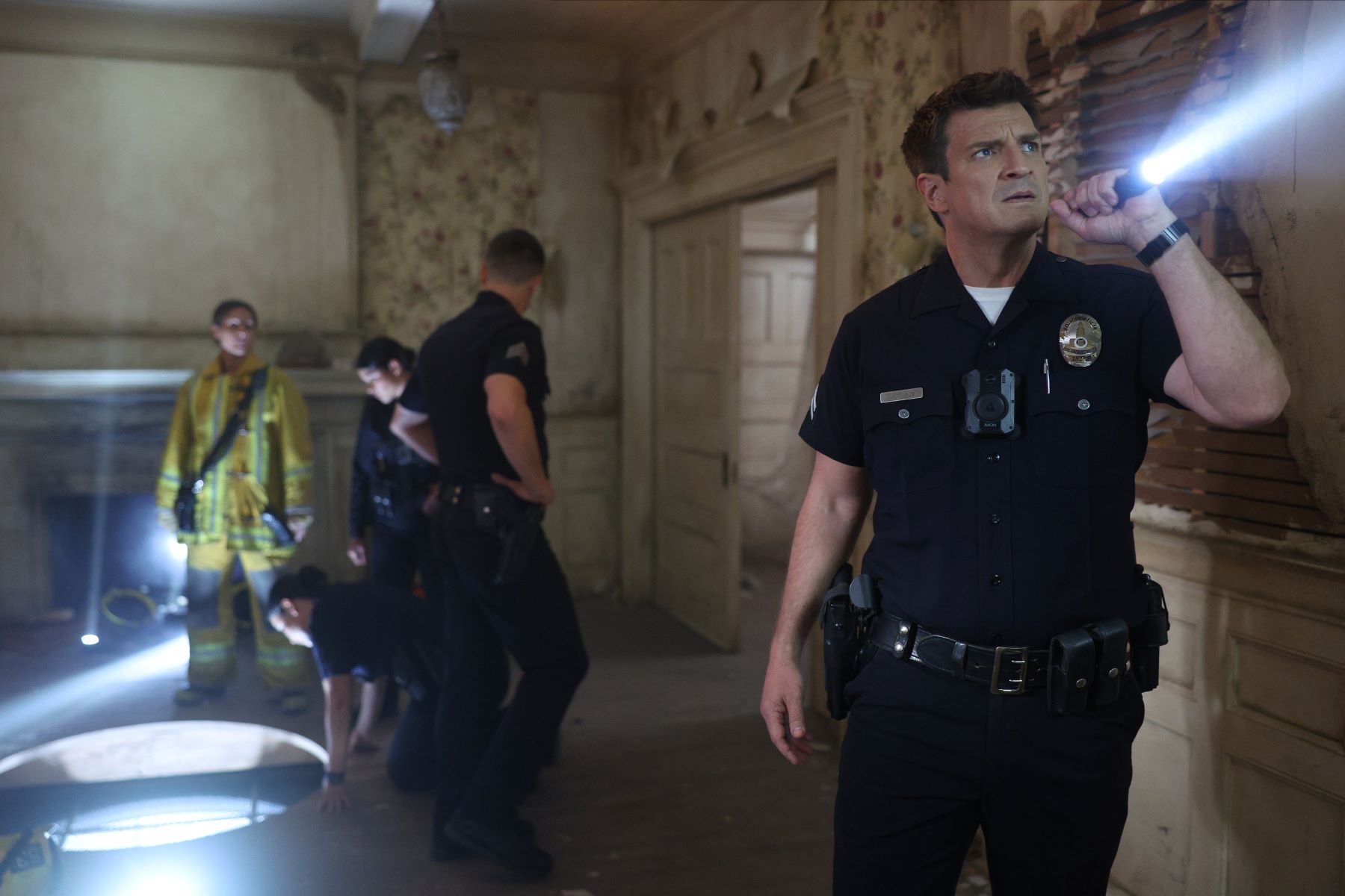 Nathan Fillion, in character as John Nolan in 'The Rookie' Season 5 on ABC, wears his dark blue police uniform while holding a flashlight.