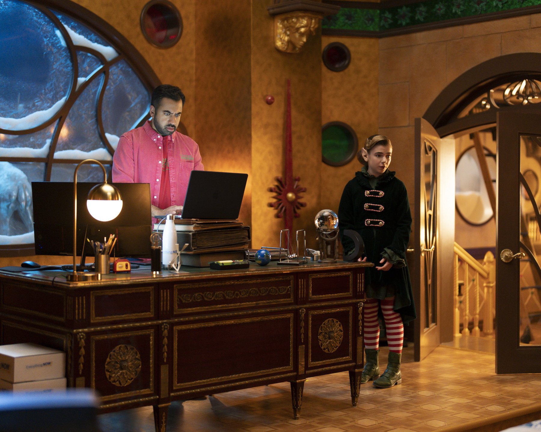 Kal Penn and Matilda Lawler as Simon Choksy and Betty in 'The Santa Clauses' for our article about episode 5. Simon is standing at his desk, looking at a computer. Betty is standing next to it,