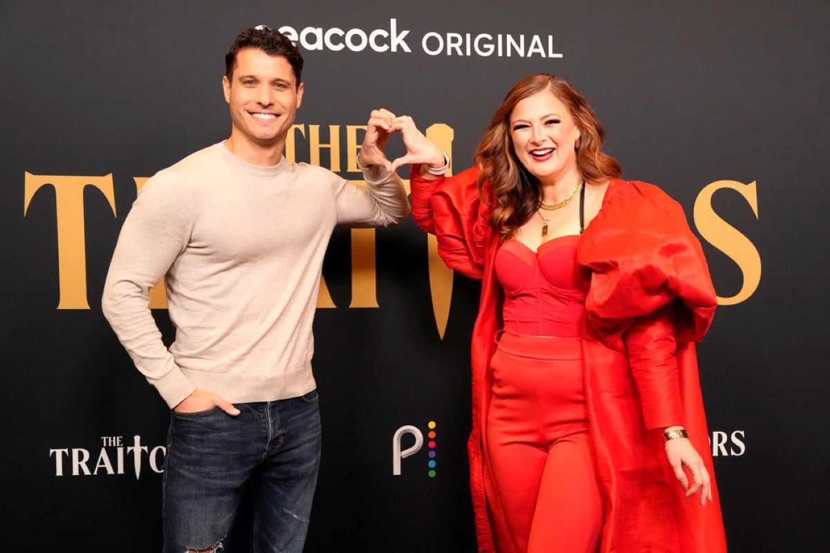 Cody Calafiore and Rachel Reilly, who are in 'The Traitors' cast, pose for pictures on the red carpet. Cody wears a cream colored sweater and jeans. Rachel wears a red jumpsuit and big red jacket.