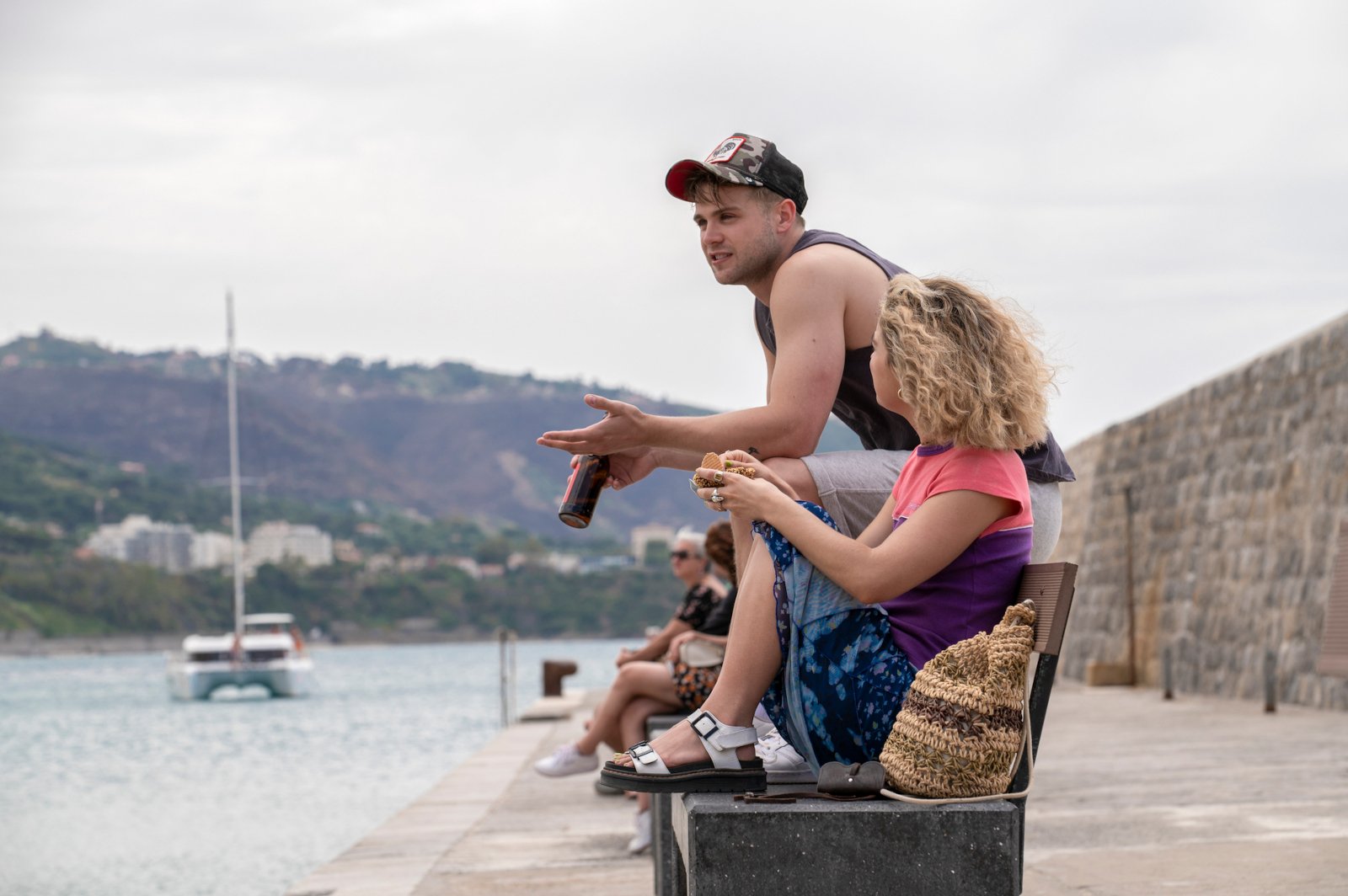 Leo Woodall and Haley Lu Richardson in 'The White Lotus' Season 2 for our article about finale theories. They're sitting on a bench in front of the water.