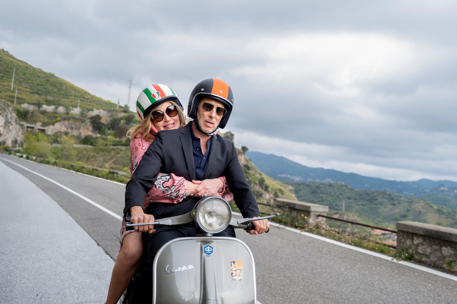 Jennifer Coolidge and Jon Gries as Tanya and Greg in 'The White Lotus' Season 2 for our article about characters who should return in season 3. They're riding a scooter with mountains in the background.