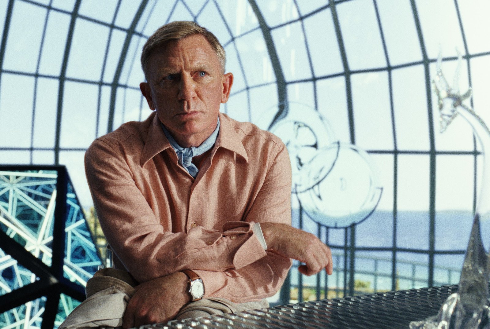 Daniel Craig as Detective Benoit Blanc in 'Glass Onion' for our article about what time it is out on Netflix. He's wearing a pink packet and in a room with glass walls.