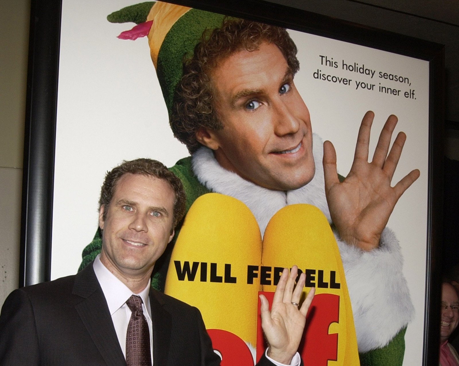 Will Ferrell standing in front of an 'Elf' poster for our article about where to stream the film in 2022. He's smiling and waving.