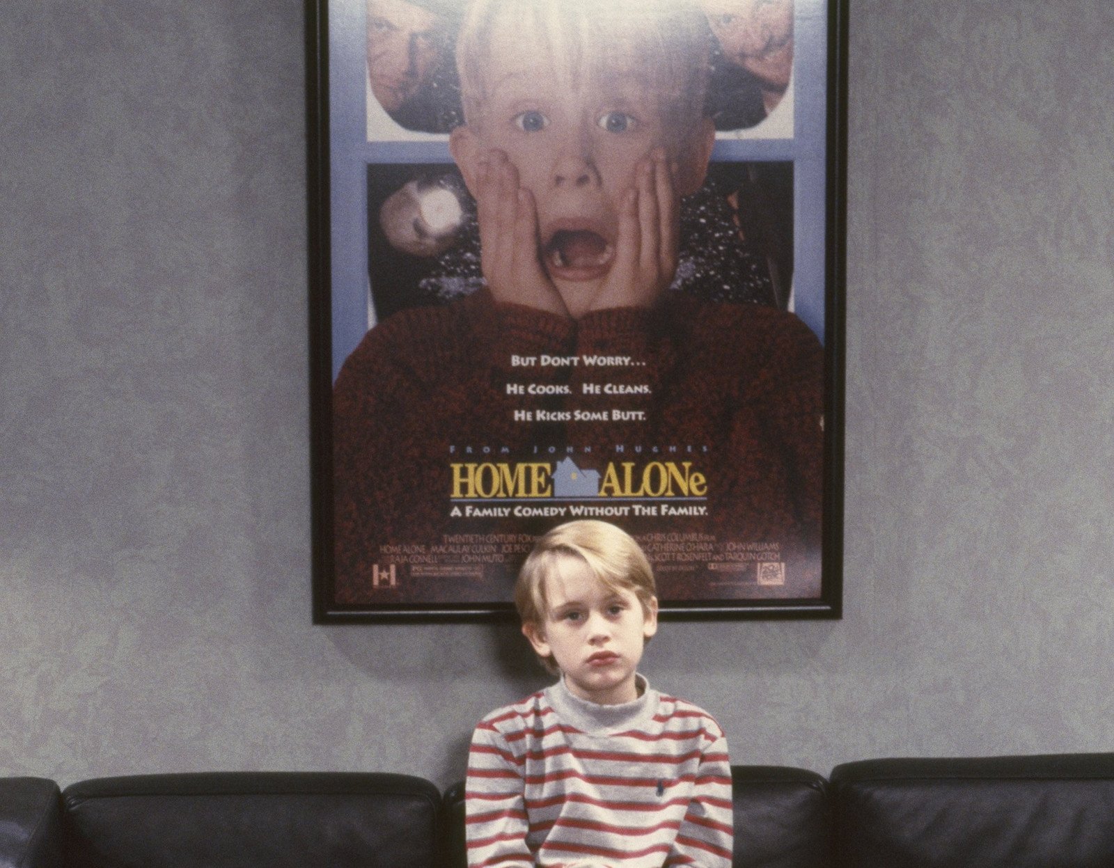 Macaulay Culkin sitting in front of a 'Home Alone' poster for our article about where to stream the movies. He's wearing a white and red striped shirt and frowning at the camera.