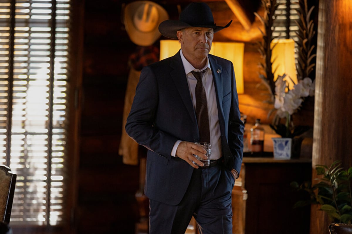 In Yellowstone, John Dutton stands in his living room holding a glass of whiskey. He is wearing a suit and tie and a black cowboy hat.