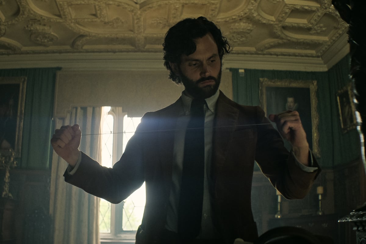 Penn Badgley as Joe Goldberg in 'You' Season 4. Joe wears a suit and tie, has a beard and longer hair, and stretches out a piece of string.