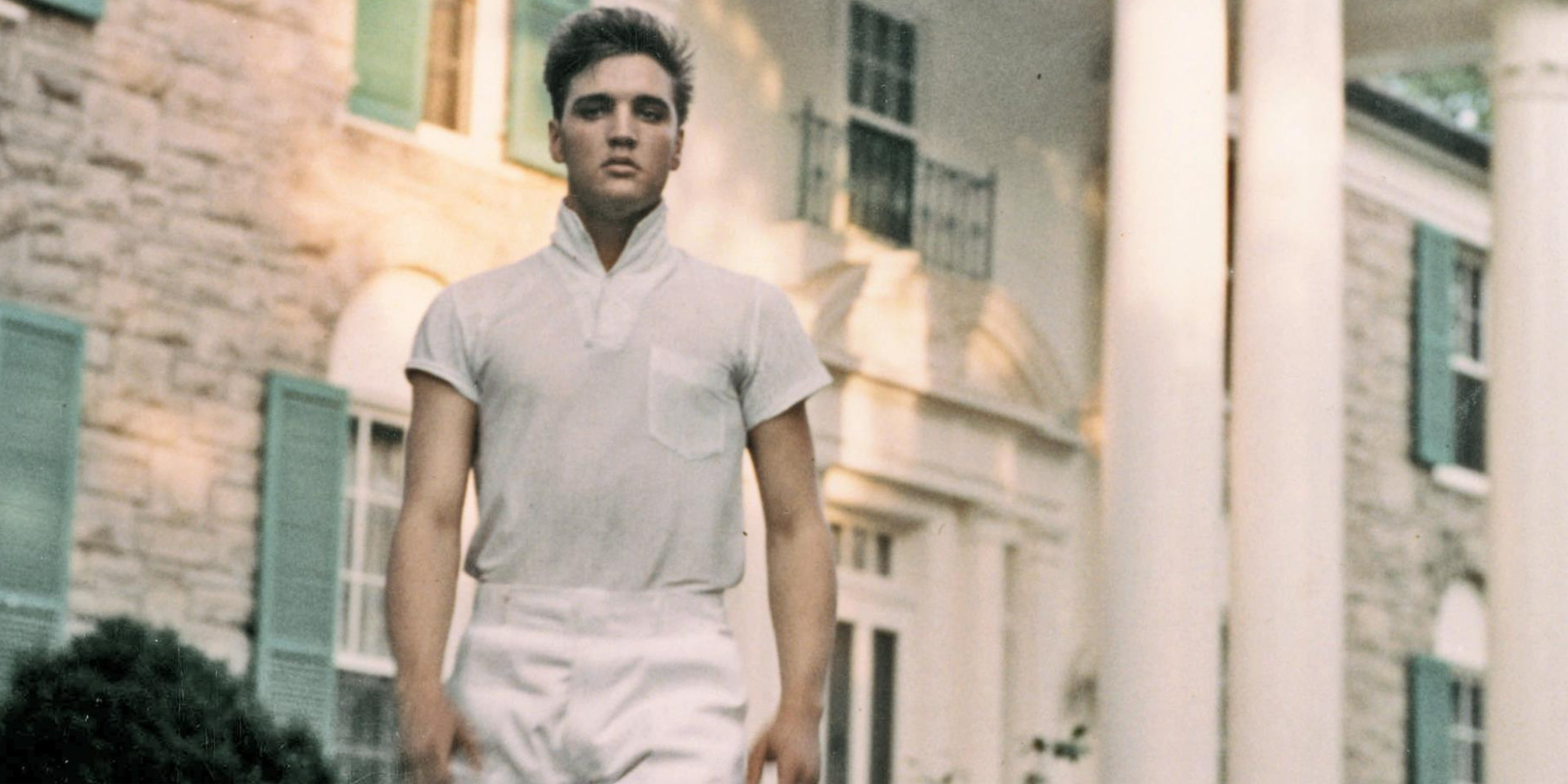 Elvis Presley poses in front of Graceland in a color photograph.