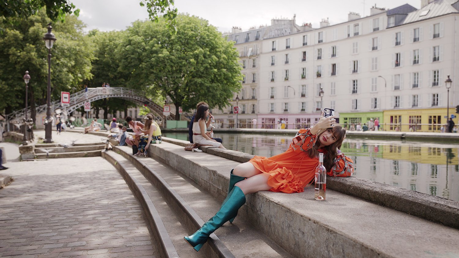 Emily in Paris cast member Lily Collins as Emily Cooper leaning back on a set of steps beside a river in season 3.