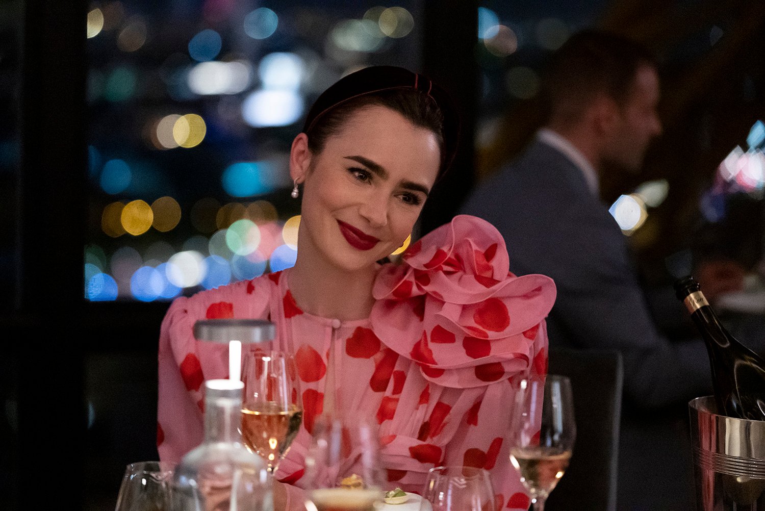 Emily in Paris Season 3: Lily Collins as Emily Cooper sitting at a dinner table surrounded by wine glasses and smiling as she wears a pink and red ruffled dress.
