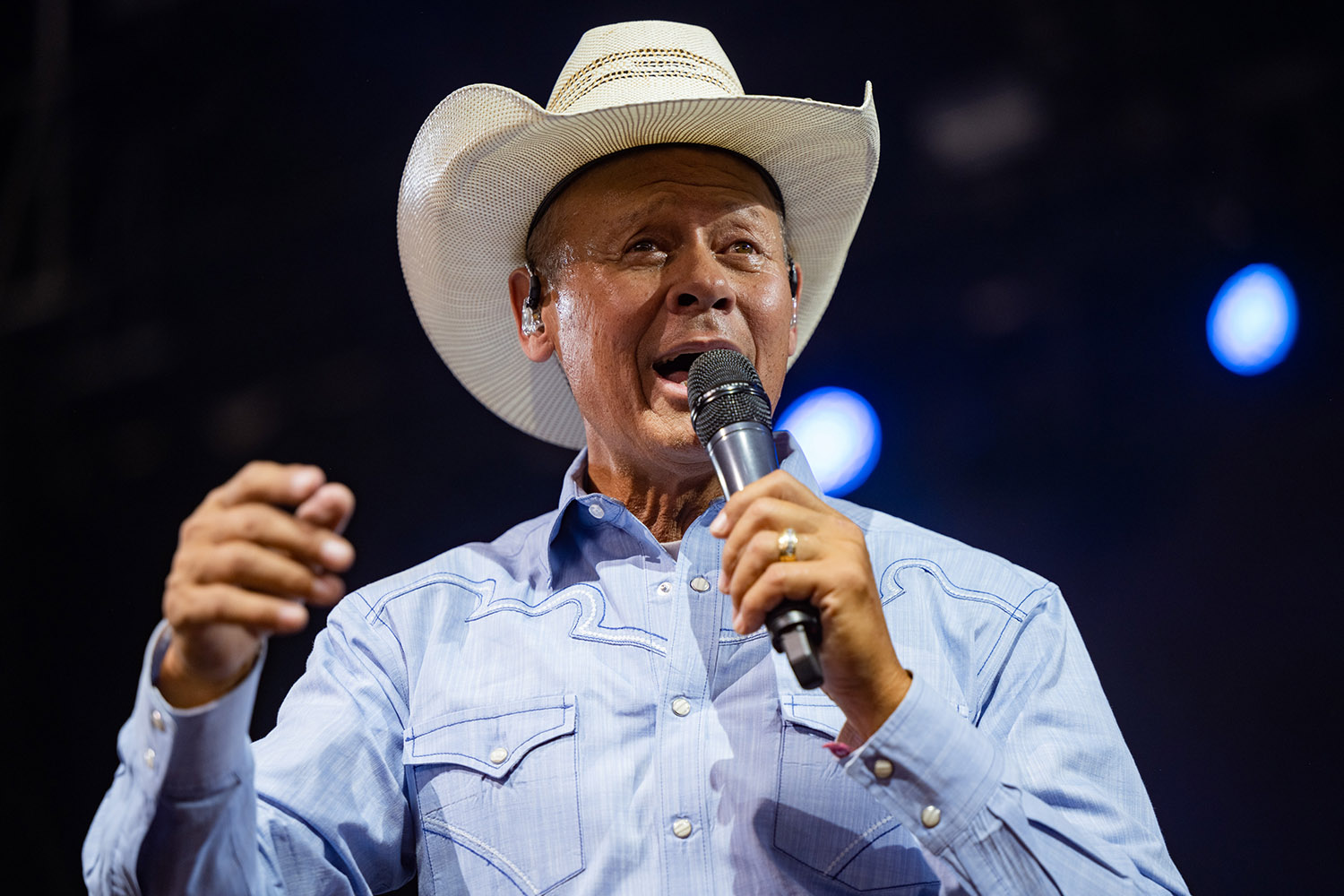Neal McCoy, whom Blake Shelton wants to replace him on The Voice, sings into a microphone while wearing a blue button-down and a cowboy hat at Stagecoach 2022