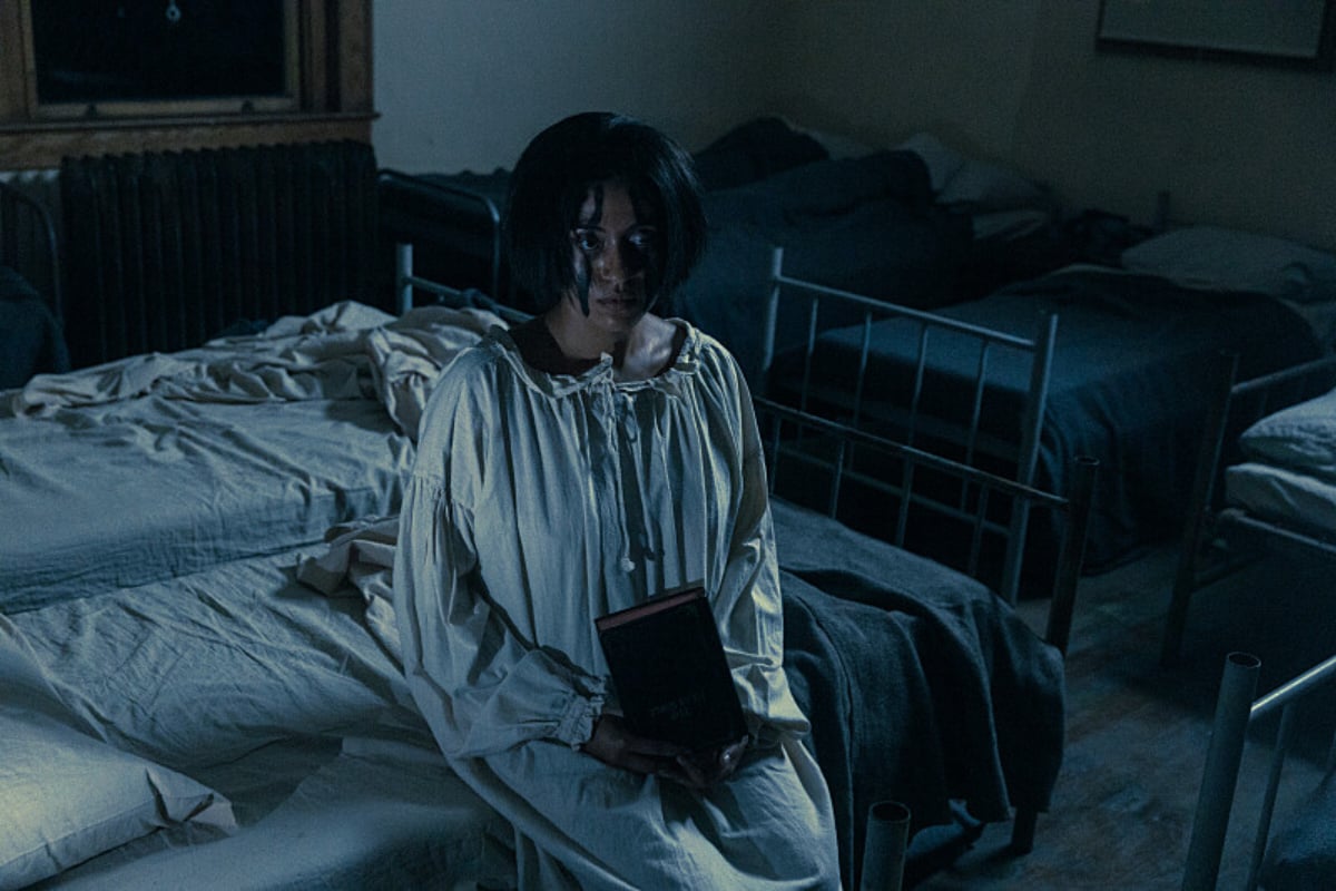 Aminah Nieves plays Teonna in 1923. Teonna sits on her bed wearing a white night gown and holding a Bible. She has short hair and black markings on her face. 