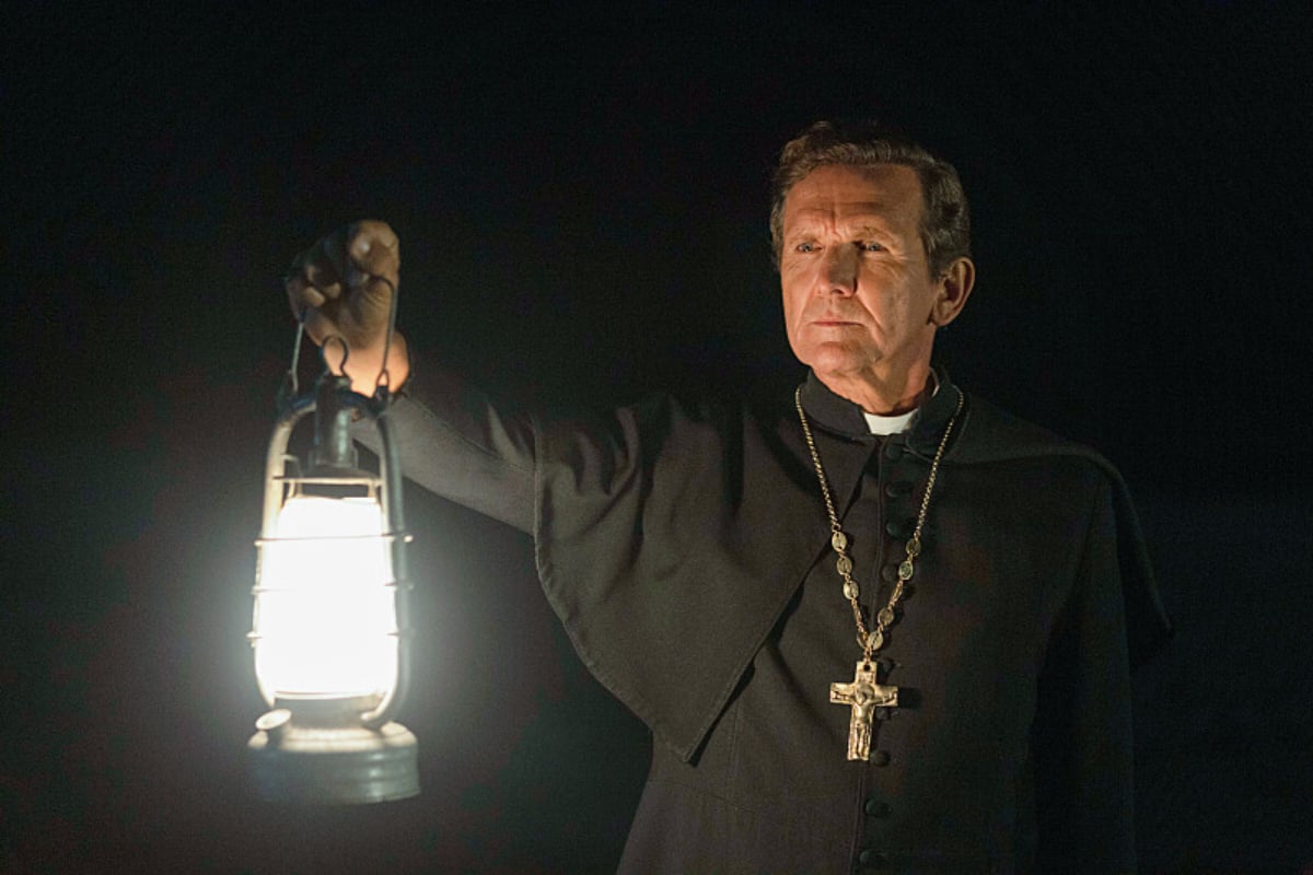 Sebastian Roché plays Father Renaud in 1923. Father Renaud wears a cassock and crucifix and carries a lantern.