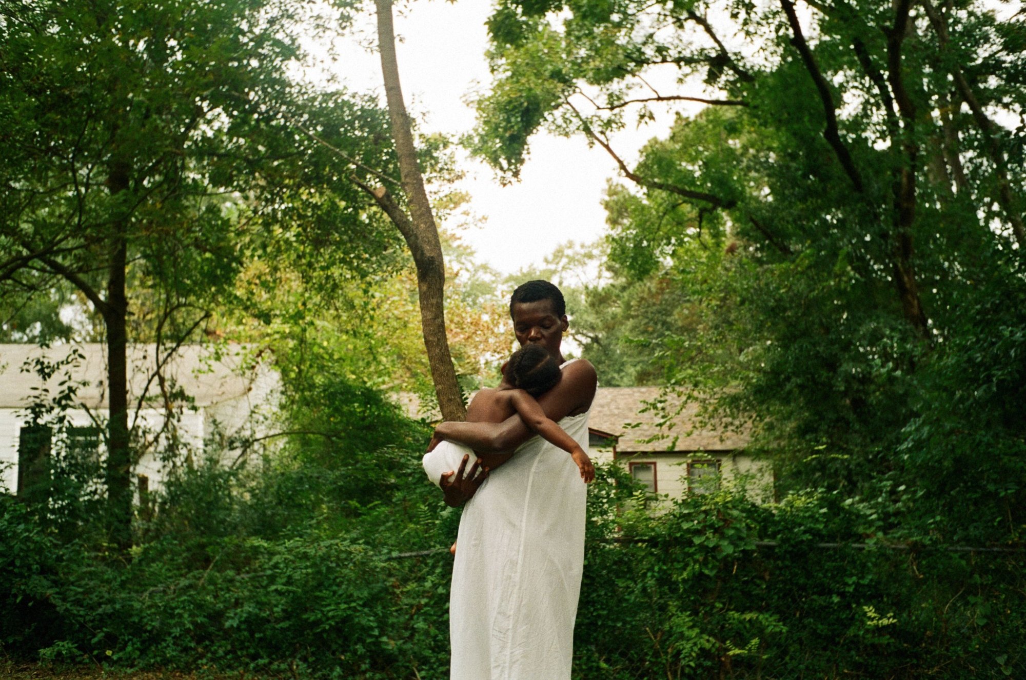 'All Dirt Roads Taste of Salt' Sheila Atim as Evelyn wearing all white, wearing a baby surrounded by greenery