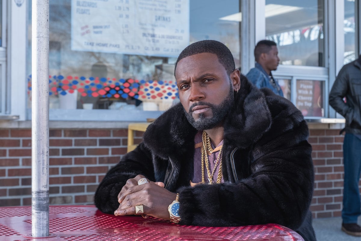 Rayan Lawrence wearing black fur and gold jewelry while sitting at a table as K-9 in 'BMF'