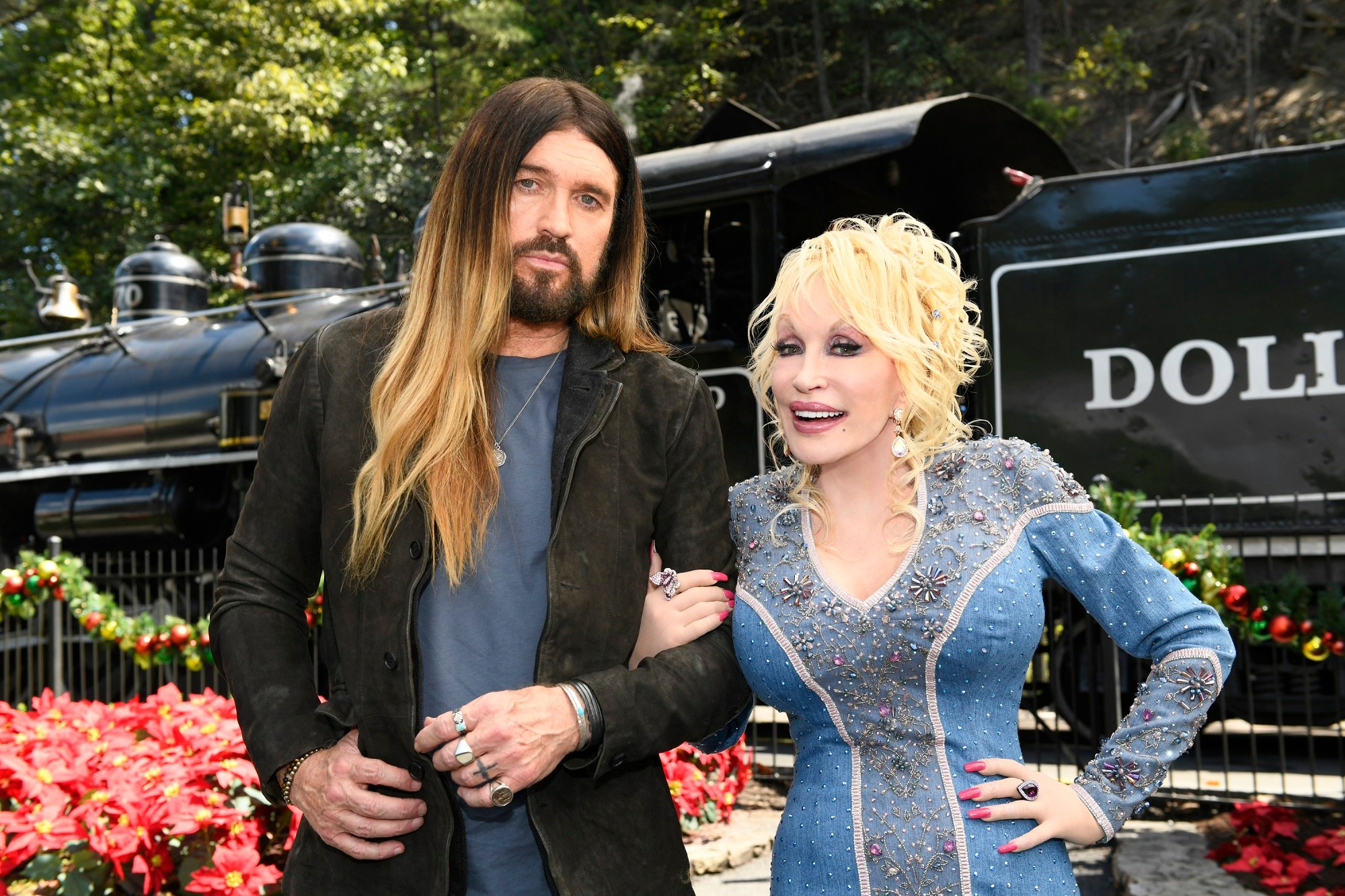 Billy Ray Cyrus and Dolly Parton stand arm-in-arm and look at the camera